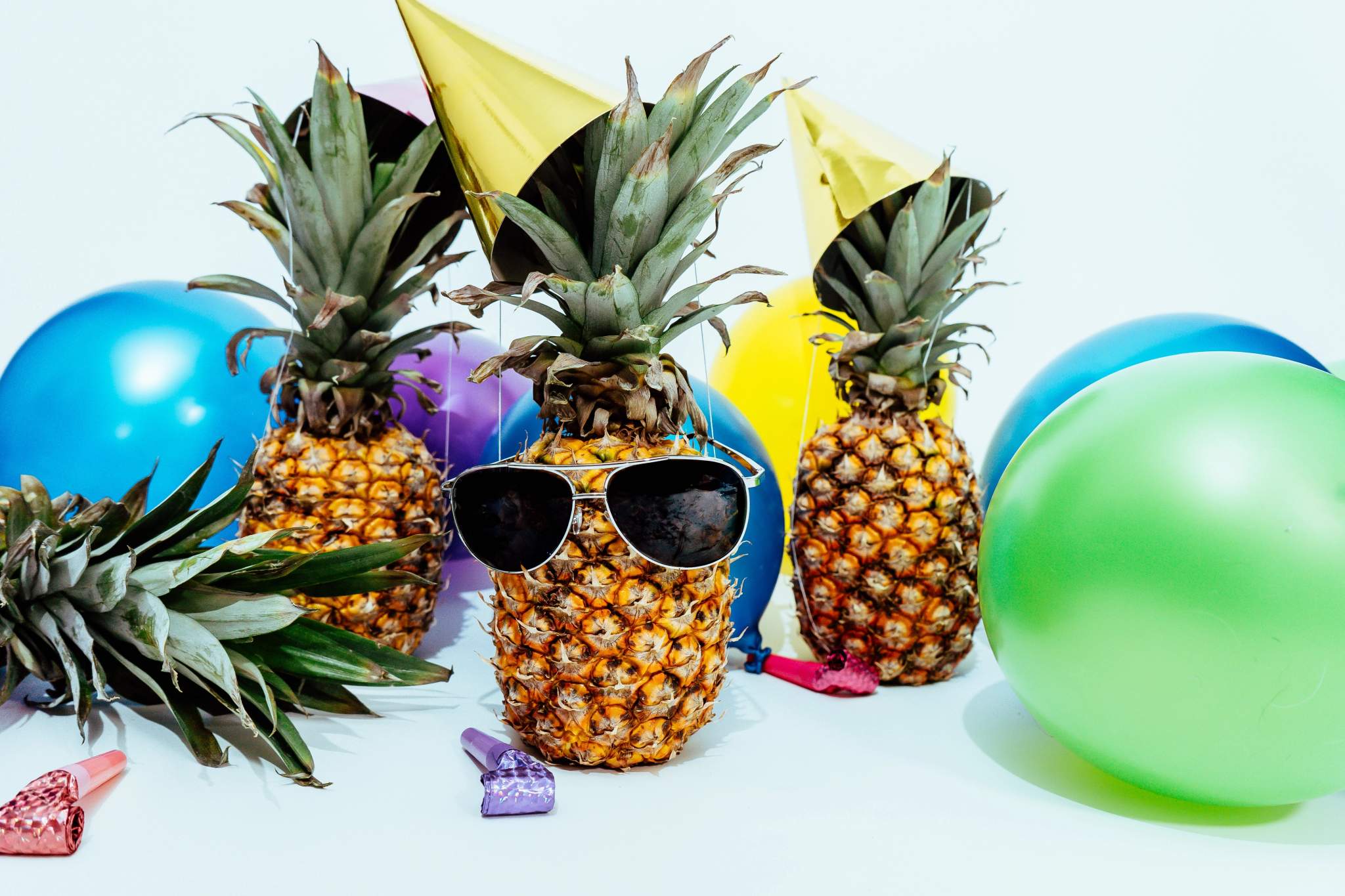 Party-hatted pineapples stand beside colourful balloons and scattered decorations.