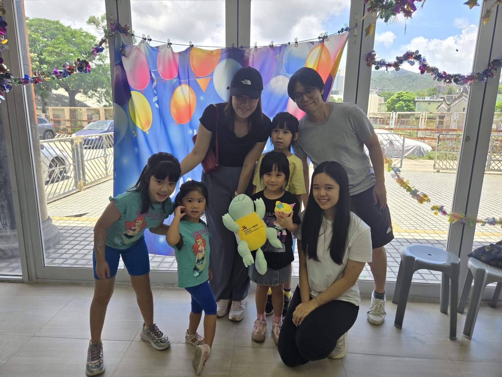Children and adults pose together with balloons and stuffed toys.