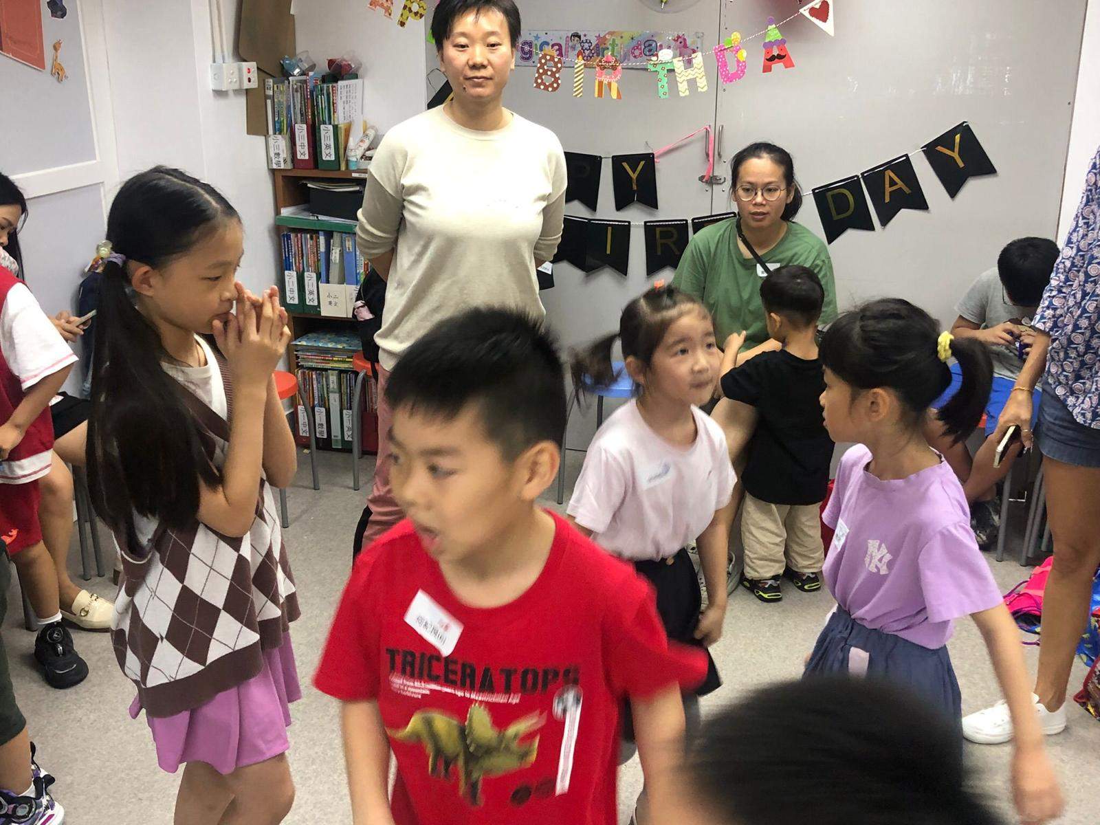 Children mingle with adults beneath colourful birthday bunting indoors.