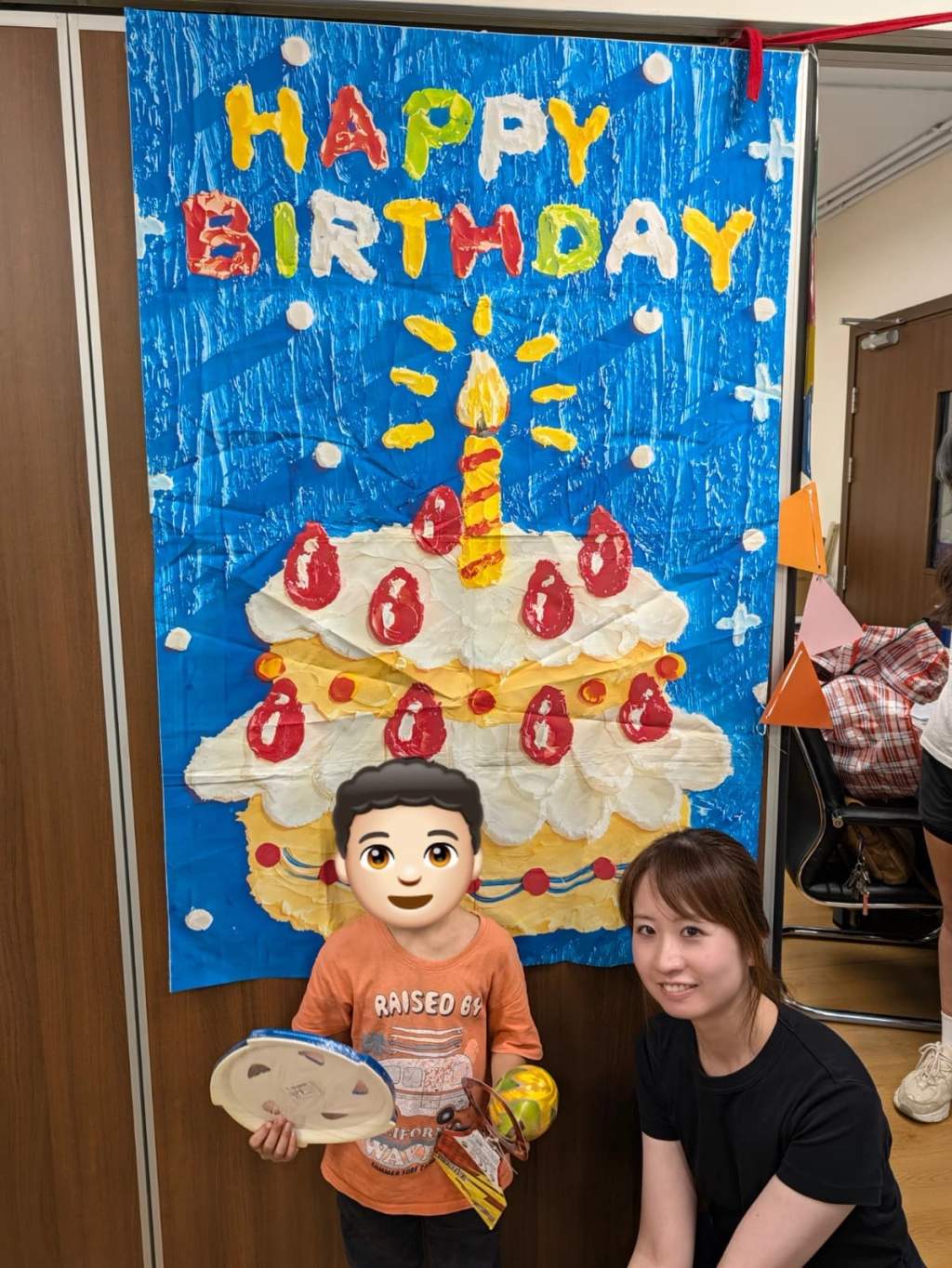 A child displays party gifts beside a volunteer and birthday backdrop.