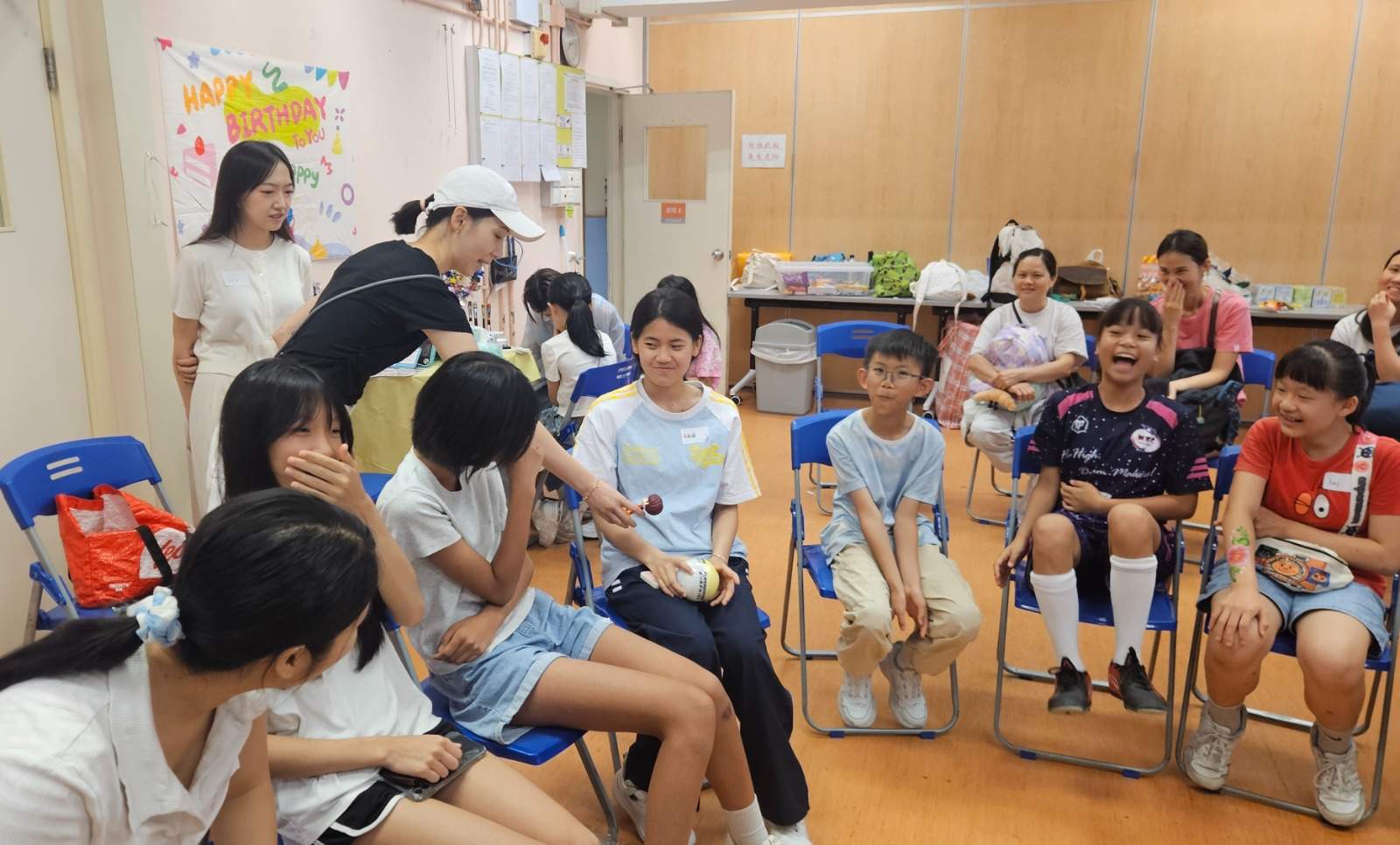 Children and adults sit in a circle on blue chairs.