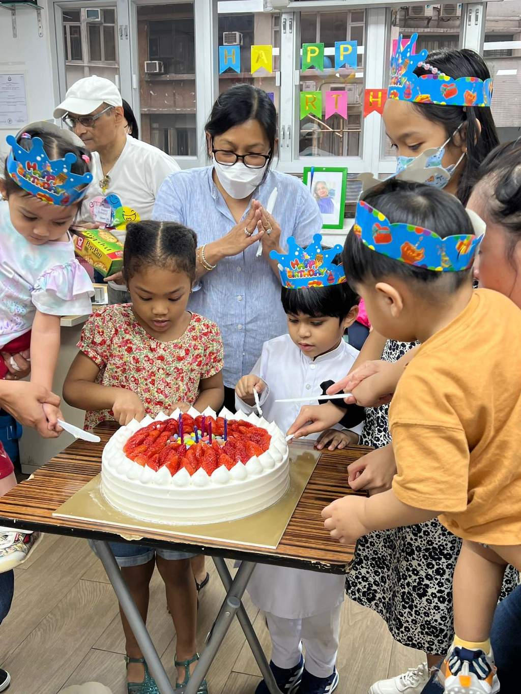Children and adults gather around a strawberry-topped cake on a table.