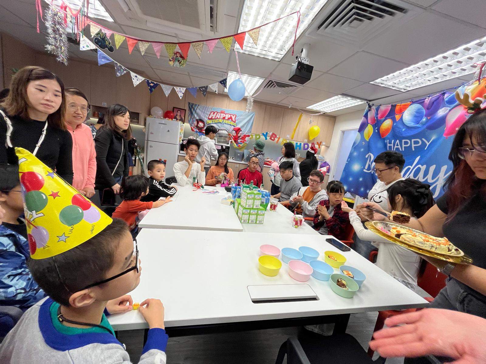 People sit along both sides of a table with party supplies.