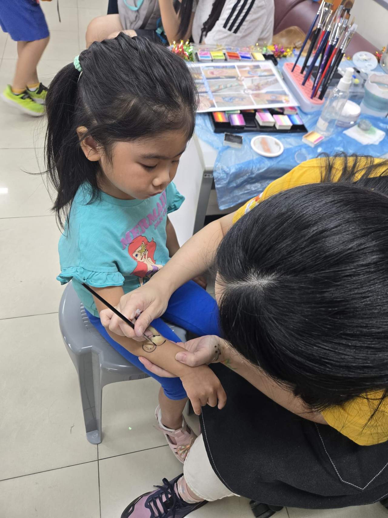 An adult paints a seated child's hand beside art supplies.