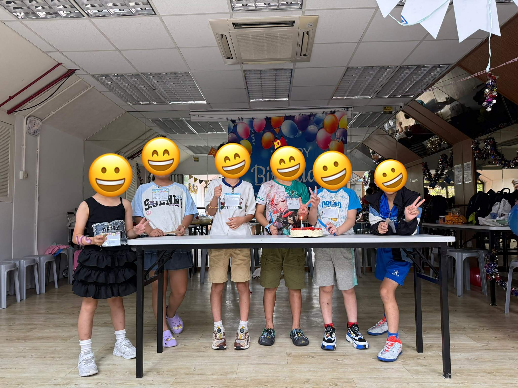 Children stand behind a cake table beneath a balloon backdrop.