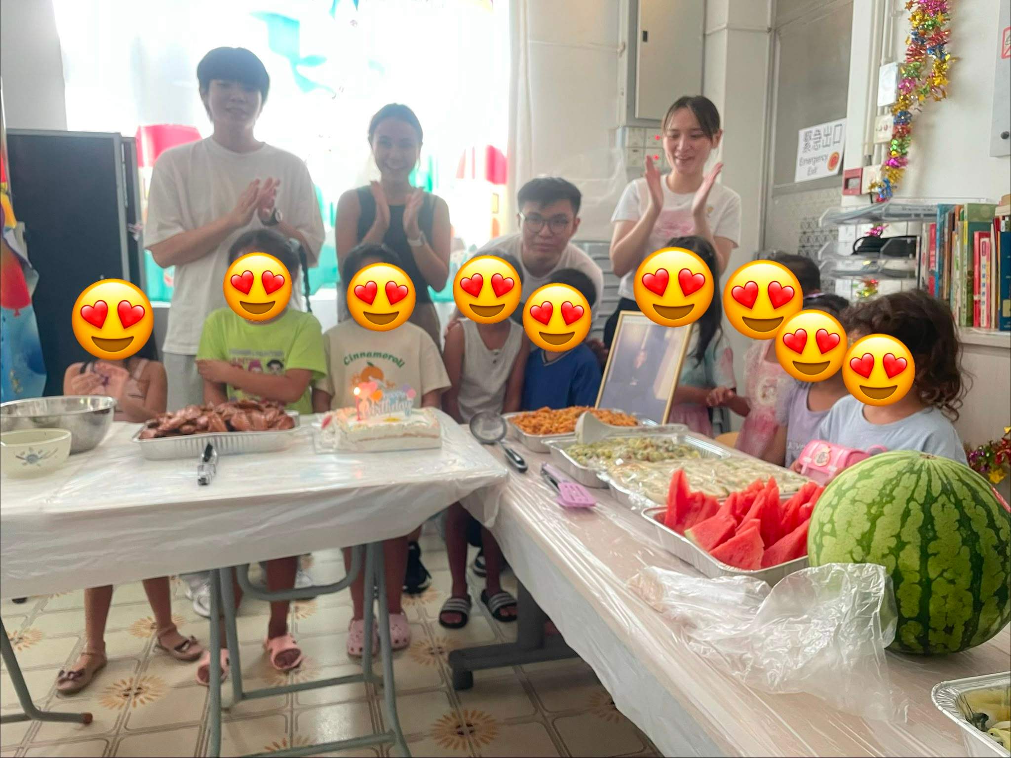 Children and adults stand behind cakes and trays of food.