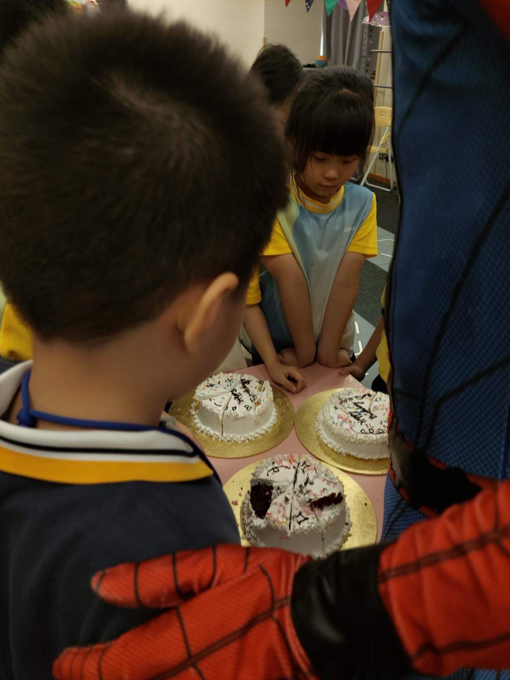 Children gather closely around three decorated cakes on a table.