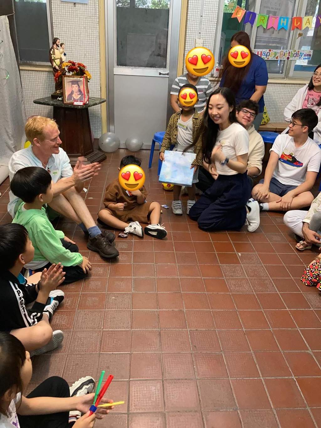 Adults and children gather in a circle around a seated child.