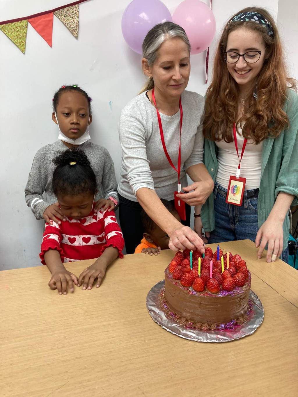 Two adults cut a strawberry-topped cake as children stand nearby.