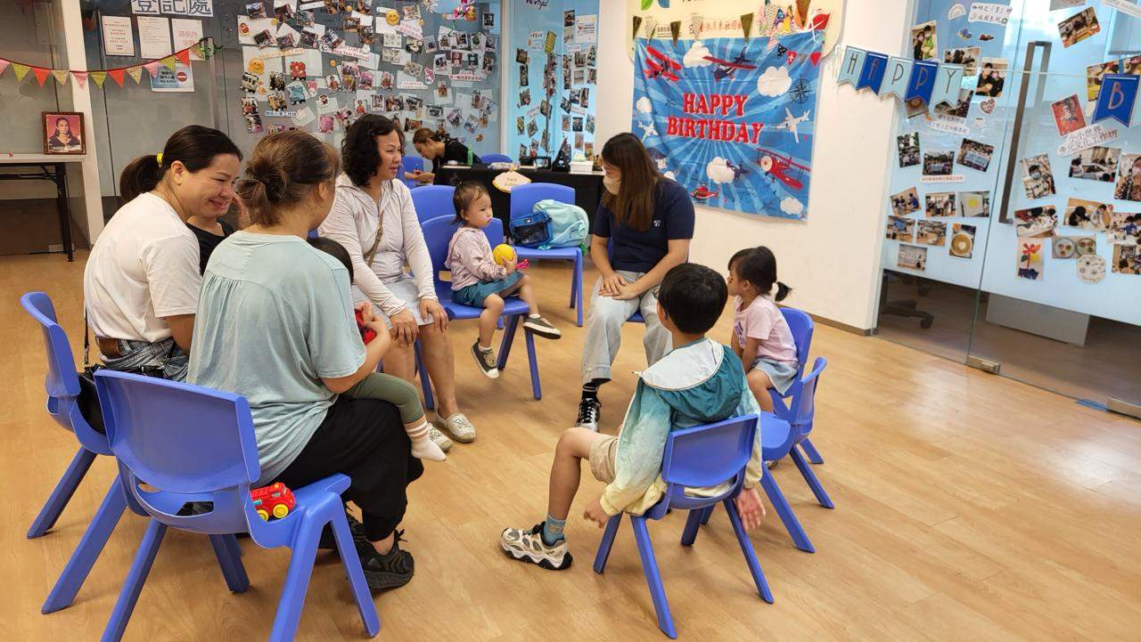 Adults and children sit together in a circle of chairs.