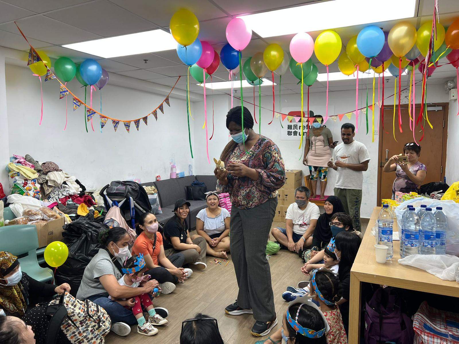 An adult stands before people seated beneath colourful balloons and bunting.
