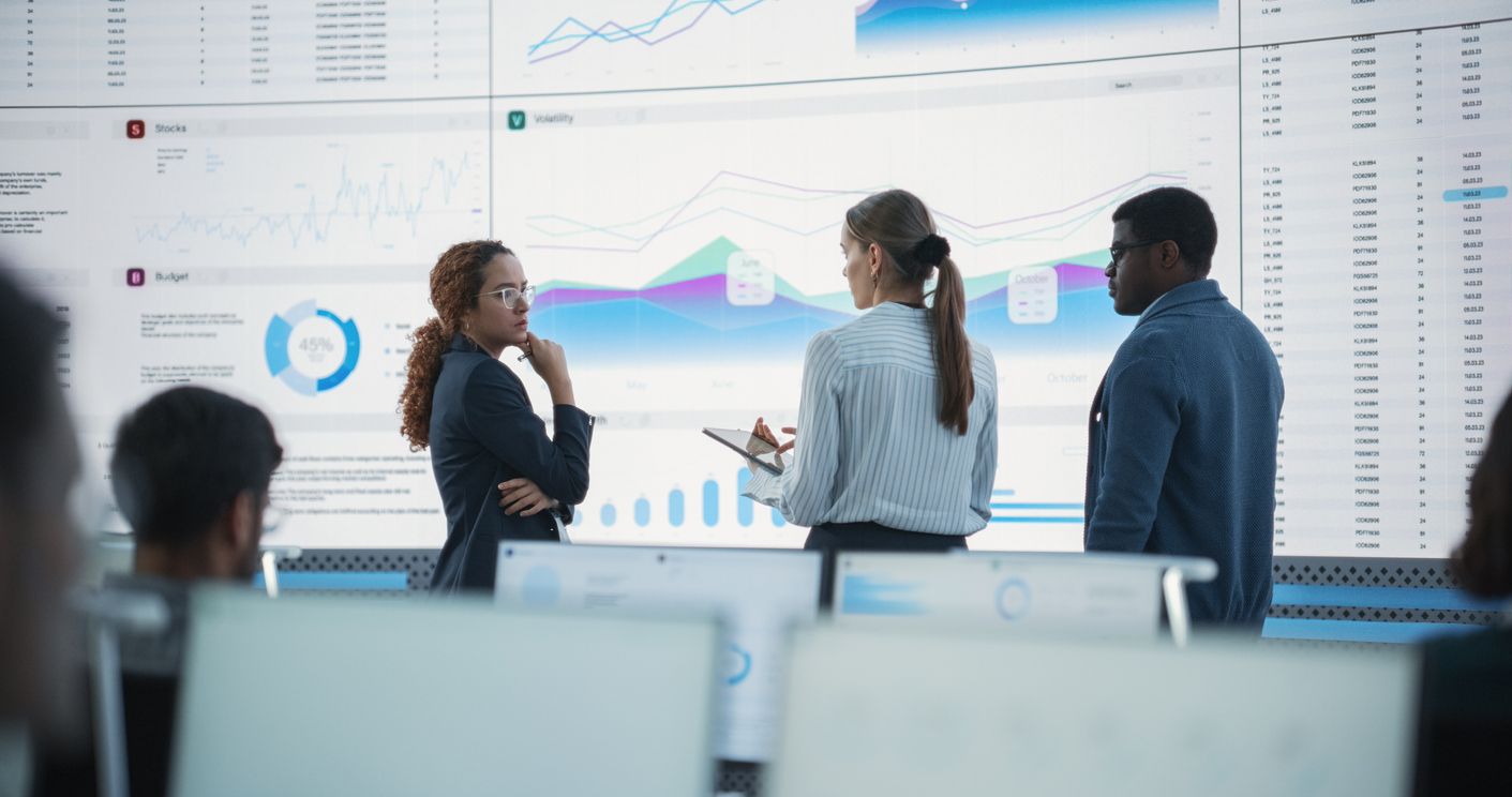 Three professionals in a modern control room analyze data displayed on large digital screens, discussing trends and insights.