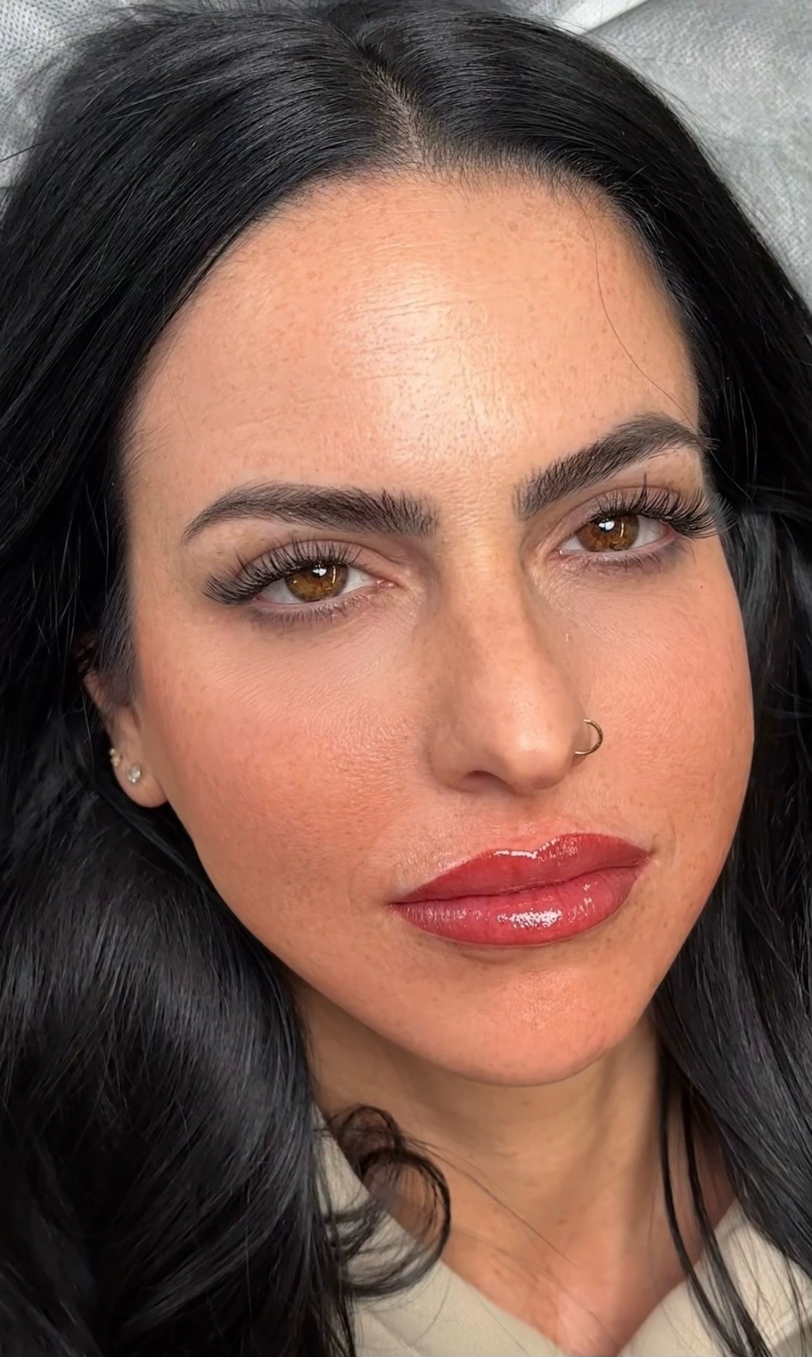 Close-up portrait of a woman with olive skin and long black hair lying back on a treatment bed, showcasing freshly completed permanent lip blush in a rich berry-red tone with a glossy finish. Her brows are well-defined with a soft, natural shape, and she has warm amber-brown eyes, full lash extensions, a small gold nose ring, and a diamond stud earring. The tight crop highlights the bold lip color and overall polished result of the permanent makeup session.