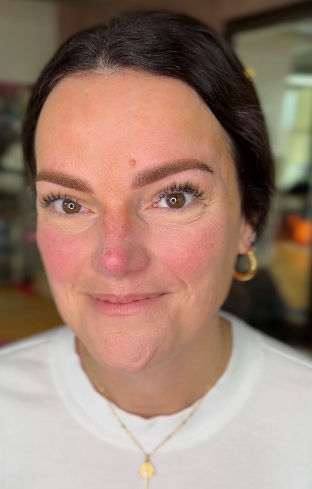 Close-up portrait of a woman with light skin and dark brown hair pulled back, smiling softly after a permanent makeup brow procedure. Her brows appear freshly done with a rich brown powder brow finish, showing defined shape and even color. She has hazel-brown eyes, lash extensions, gold hoop earrings, and a delicate gold pendant necklace over a white crew-neck top. Slight redness around the brow area indicates the procedure was recently completed.