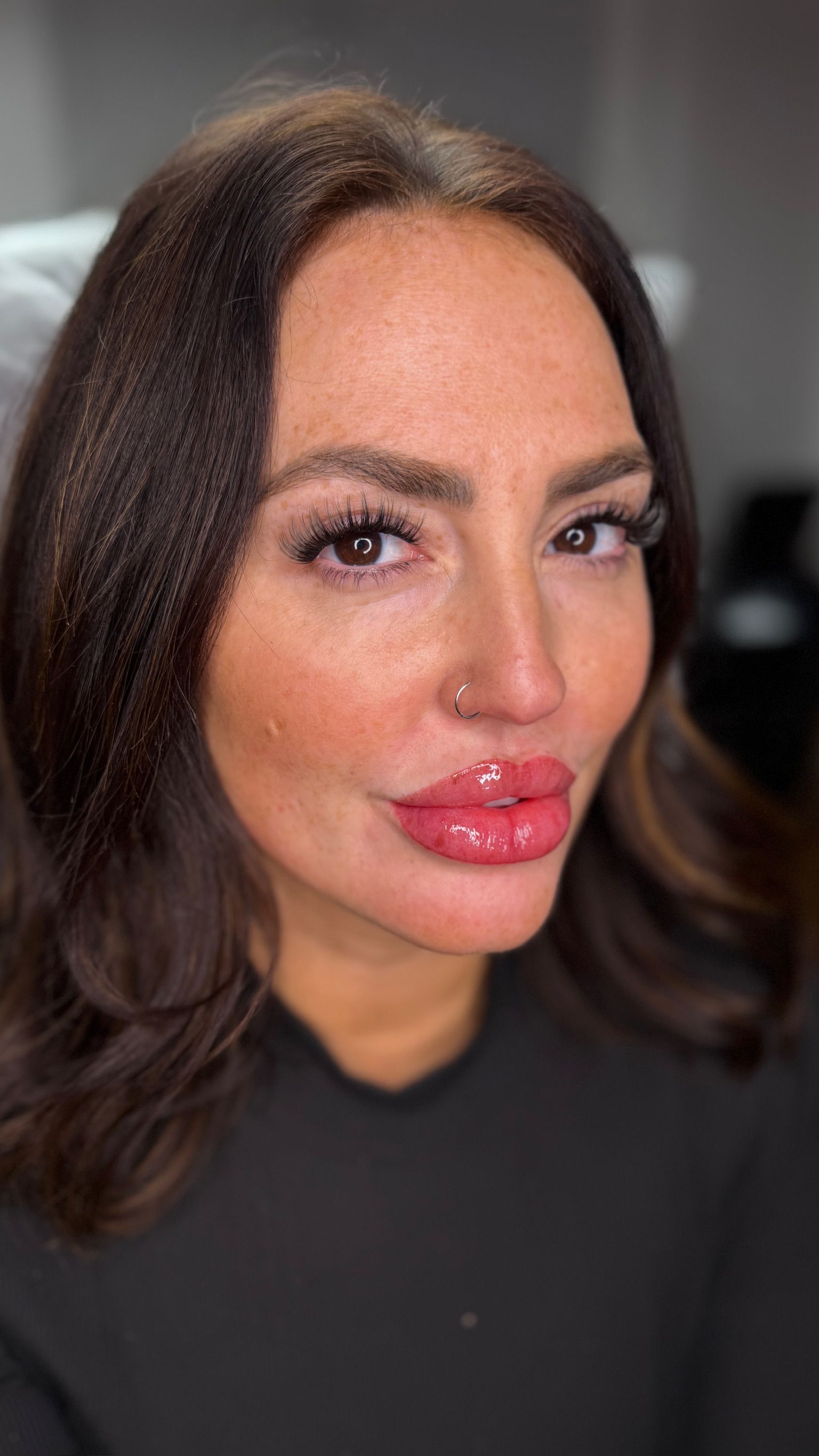 Close-up portrait of a woman with olive skin and shoulder-length dark brown hair with warm highlights, showcasing freshly applied permanent lip blush. Her lips have a vibrant, glossy rose-pink tint with a defined shape, giving a just-applied lipstick effect. She has dark brown eyes, full lash extensions, natural freckles across her cheeks and forehead, and a small silver nose ring. She wears a black top against a soft, blurred background.