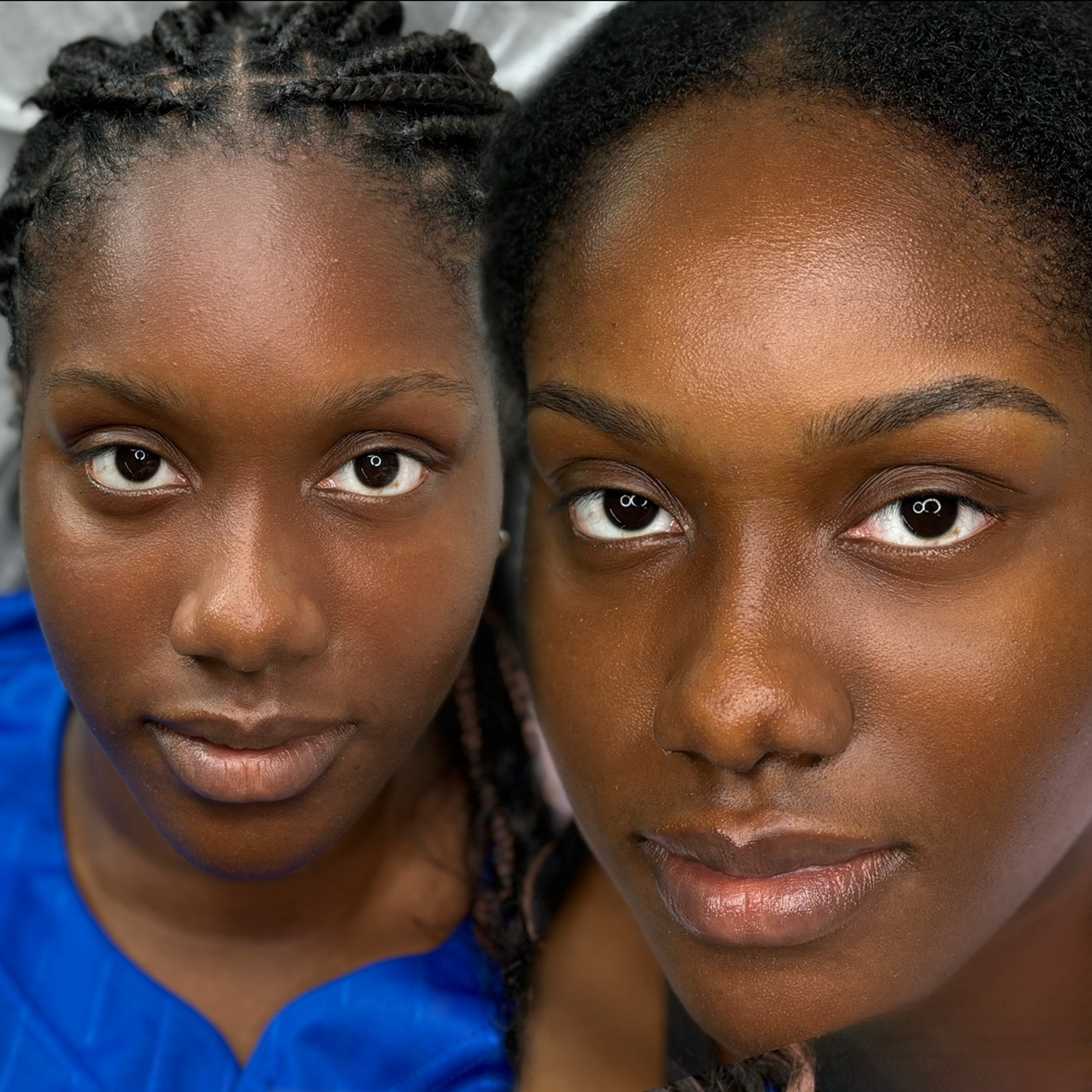 Before-and-after close-up of a young woman with deep brown skin showcasing a permanent makeup brow transformation. The left side shows her natural brows, while the right side reveals fuller, more defined brows with a clean arch. She has braided locs, dark brown eyes, and a dewy complexion. The tight crop highlights the brow enhancement and her flawless skin.