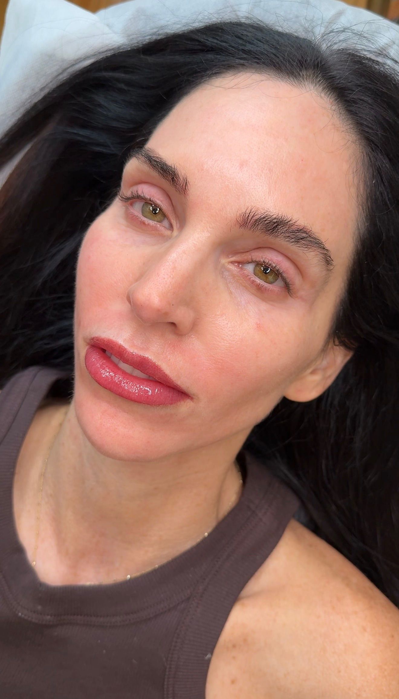 Overhead close-up of a woman with fair skin and long black hair resting on a treatment bed after a permanent makeup session. Her lips feature a fresh lip blush in a muted mauve-rose tone with a soft, natural finish. Her brows show delicate hair-stroke work with a natural, feathered shape. She has striking green-hazel eyes, minimal makeup, a delicate gold necklace, and wears a taupe-brown athletic top. The bare-faced, no-makeup look highlights how the lip blush and brow work enhance her features naturally.