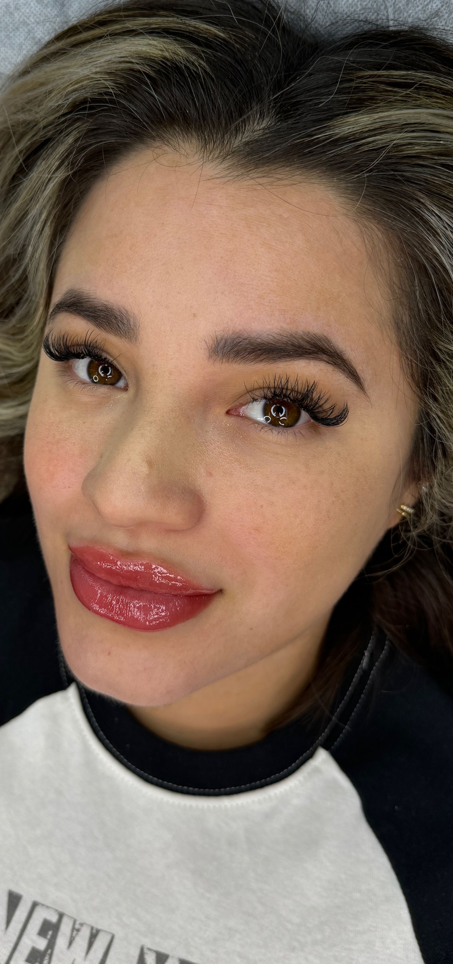 Overhead close-up of a young woman with warm-toned skin and highlighted brown-to-blonde hair lying on a treatment bed after a permanent lip blush procedure. Her lips feature a fresh lipstick-effect lip blush in a coral-pink shade with glossy balm applied, showing full, even color coverage with defined edges. She has warm brown eyes, dramatic lash extensions, softly shaped brows, a small gold earring, and wears a gray and black crew-neck sweatshirt. Her subtle smile highlights the polished, just-done result.