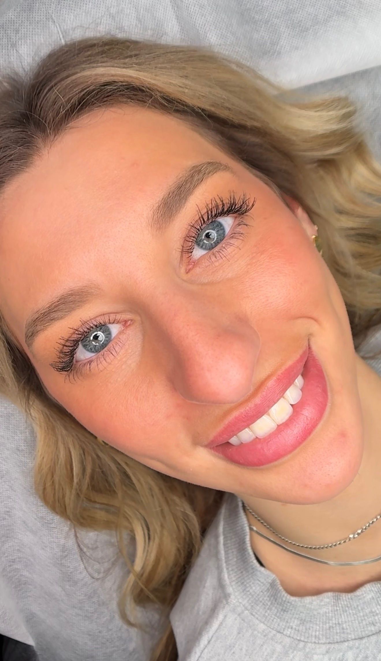 Overhead close-up of a smiling woman with light skin and wavy blonde hair lying on a treatment bed, showcasing healed lipstick-effect lip blush. Her lips display a soft, even rose-pink tone with defined edges that mimic the look of wearing lipstick, demonstrating the natural-looking results after healing. She has bright blue eyes, long lashes, softly defined brows, a small gold earring, and a silver chain necklace over a light gray top. Her wide smile highlights how the healed lip blush integrates seamlessly with her natural complexion.