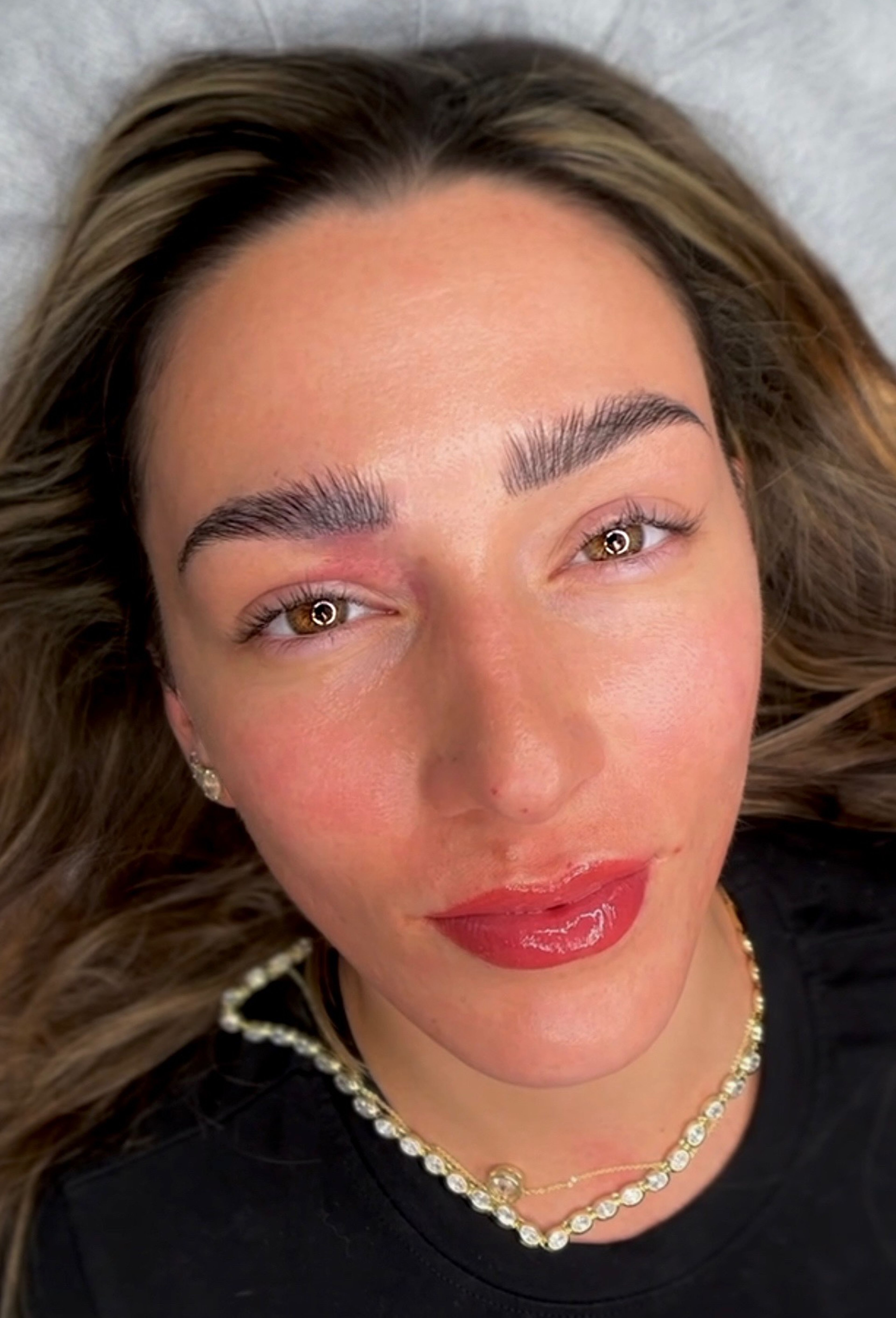 Overhead close-up of a woman with light skin and balayage brown-to-blonde hair lying on a treatment bed, showcasing freshly completed permanent makeup. Her brows feature crisp, detailed hair strokes in a dark brown tone, and her lips display a vibrant berry-red lipstick-effect lip blush with a glossy balm finish, giving the look of perfectly applied tinted lips. Slight redness around the brow area indicates the procedure was just completed. She has warm amber eyes, small diamond hoop earrings, a gold and diamond tennis necklace, and wears a black top.