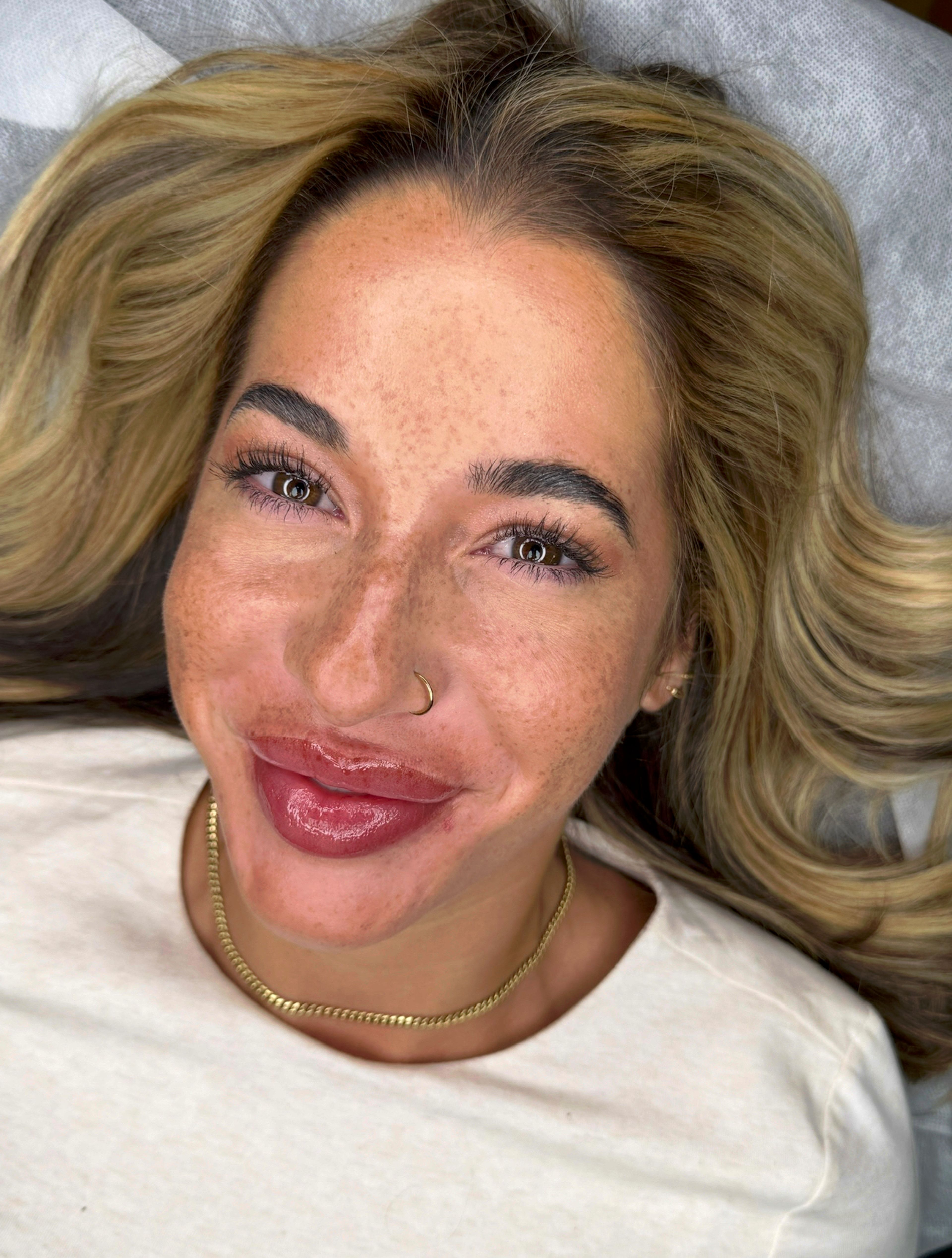 Overhead close-up of a woman with blonde hair and light, freckled skin lying back on a treatment bed, smiling after a permanent makeup procedure. Her brows show a natural, hair-stroke finish with soft definition, and her lips have a fresh rosy-pink lip blush tint with a glossy balm applied over them. She has hazel-green eyes, a small gold nose ring, a gold chain necklace, and a small stud earring. The angle and setting indicate this was taken in a permanent makeup studio immediately after the session.