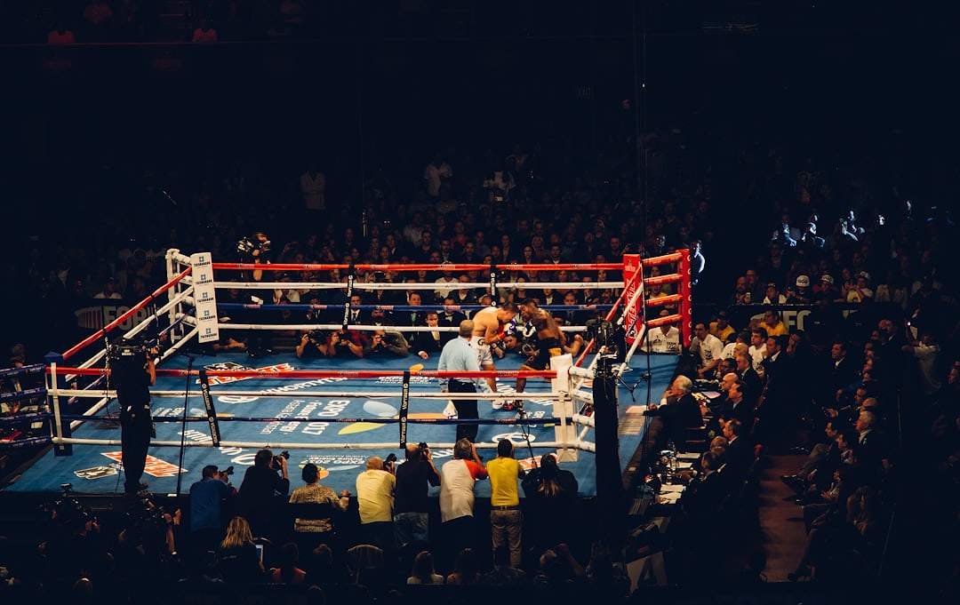 A referee and two boxers in a ring under the lights, seen from the crowd
