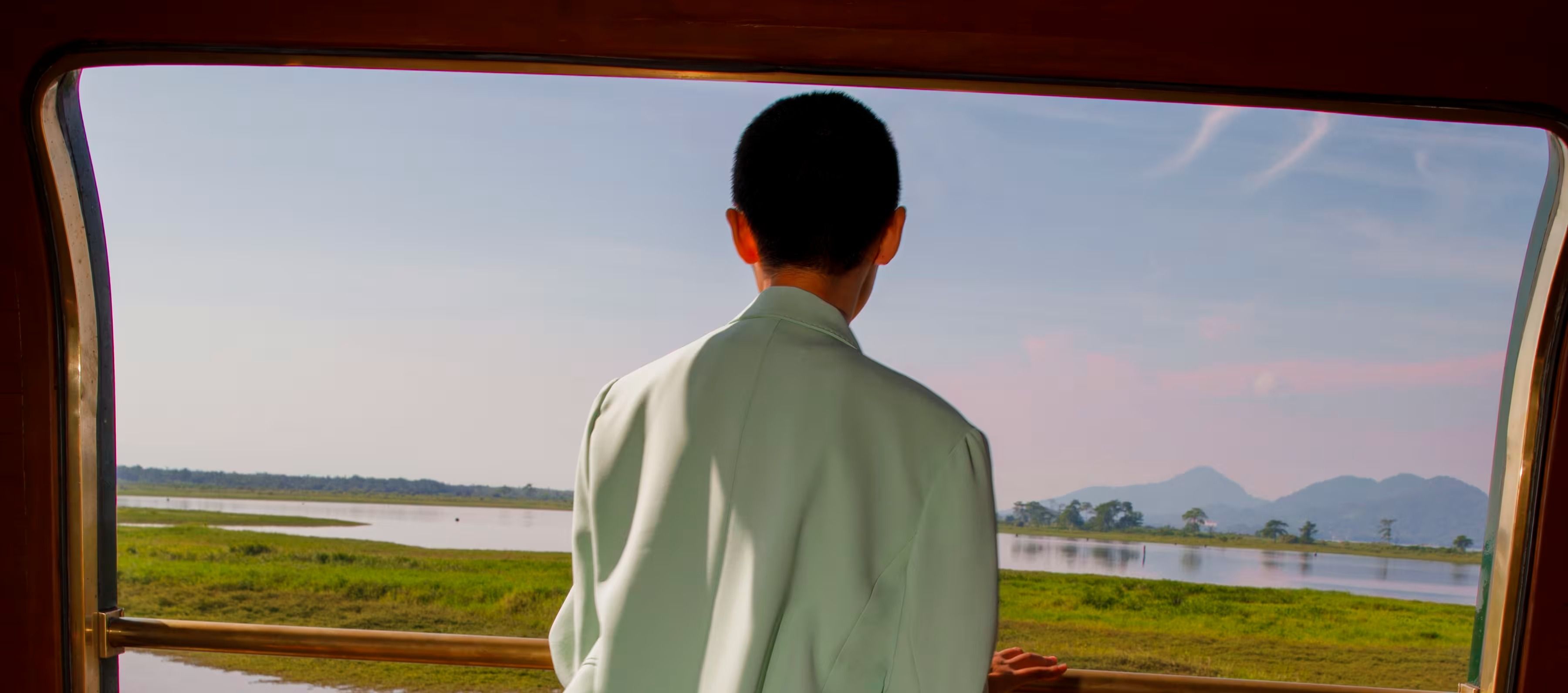 A guest at the open observation-car window of the Eastern & Oriental Express, looking out over Malaysian wetlands and distant hills.
