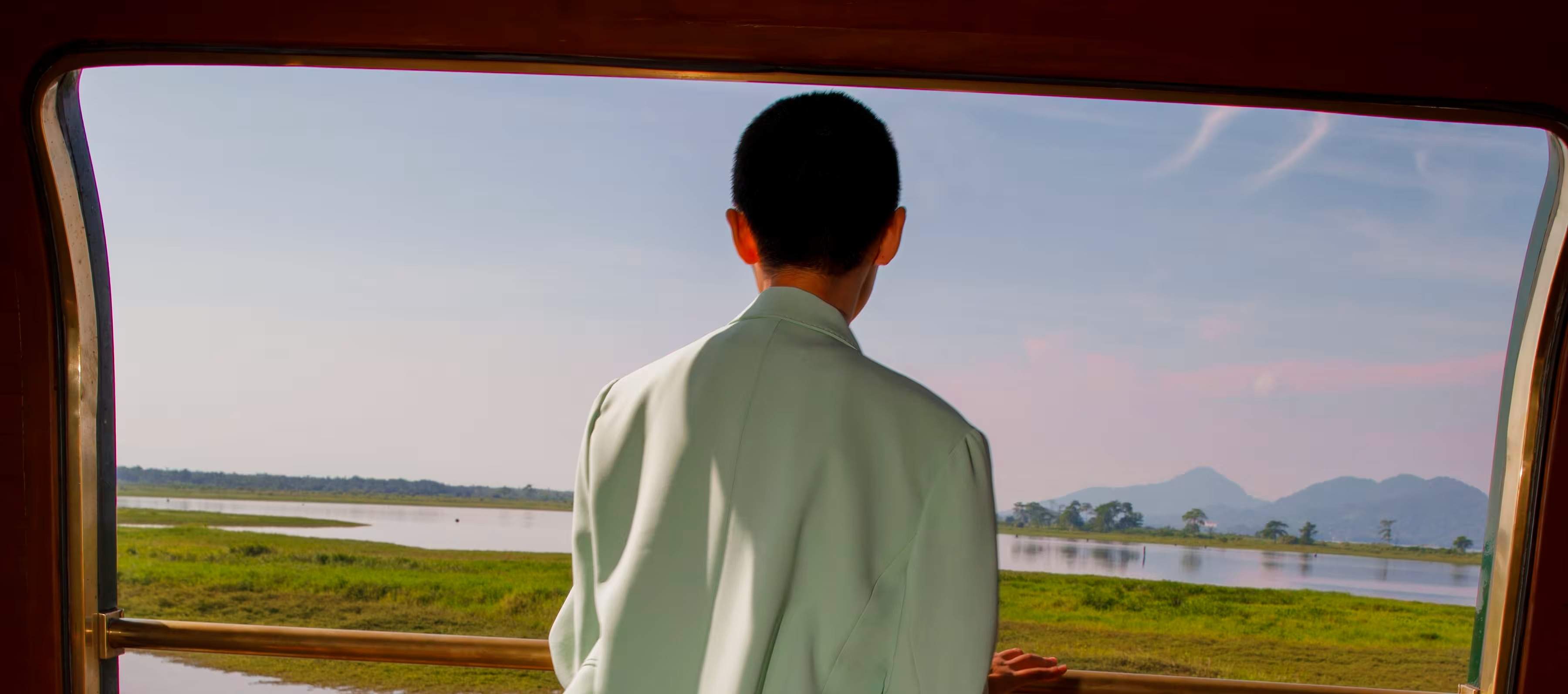A guest on the open-air observation deck of the Eastern & Oriental Express at dusk, watching Malaysian countryside pass