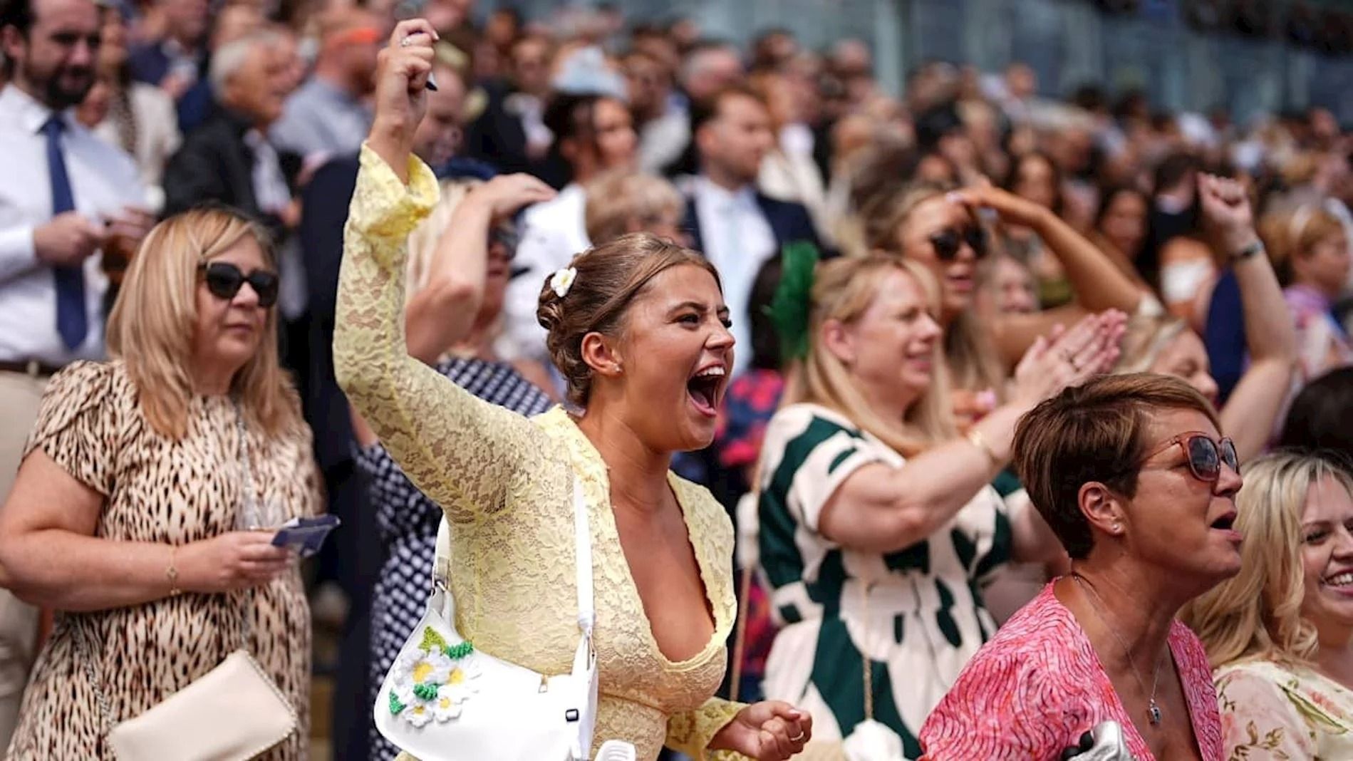 Women in the crowd at Epsom in summer dresses, a yellow lace dress, a leopard print dress and floral prints, most without hats