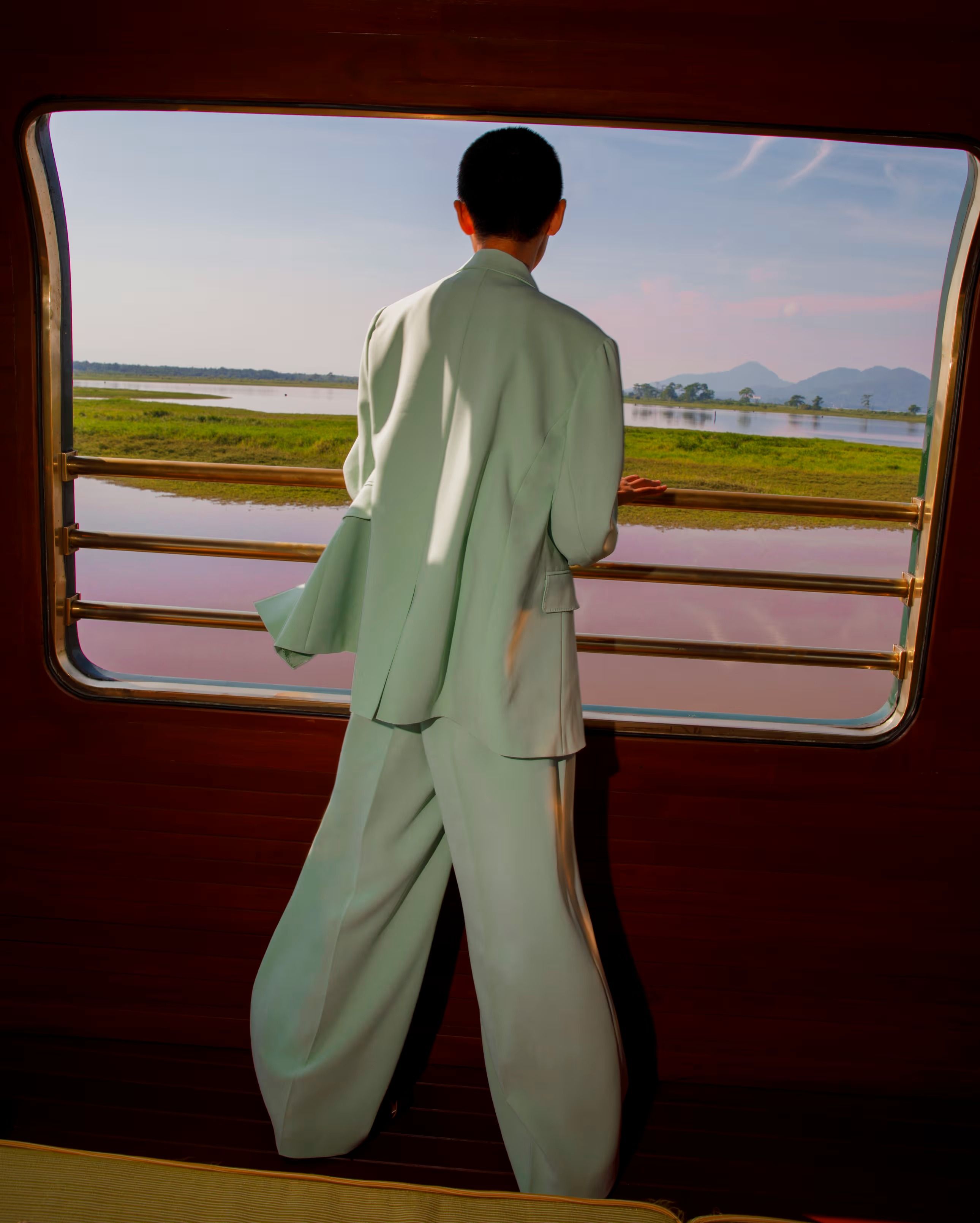 A guest at an open observation-car window of the Eastern & Oriental Express, looking out over Malaysian wetlands and distant hills.