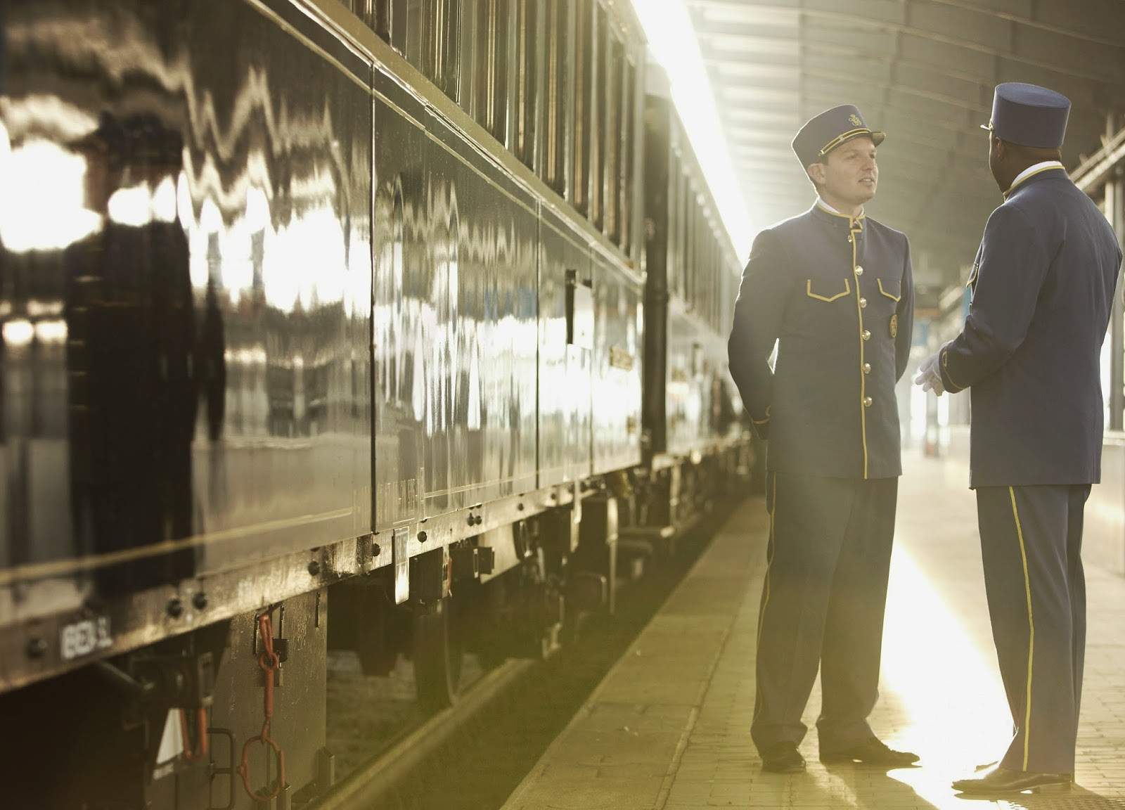 A steward greeting a guest beside the dark-blue Venice Simplon-Orient-Express at a platform in golden light