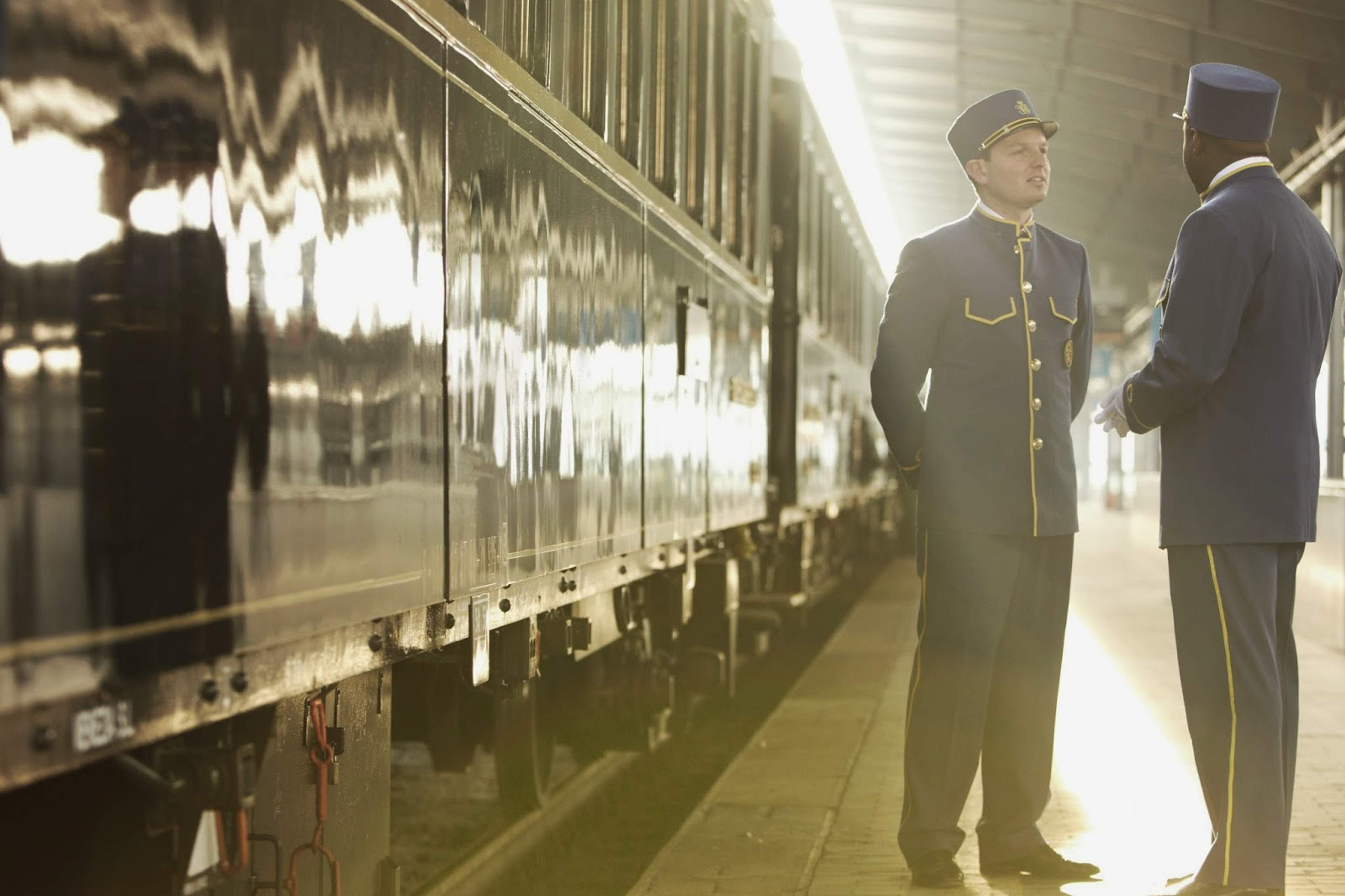 A steward greeting a guest beside the dark-blue Venice Simplon-Orient-Express at a platform in golden light