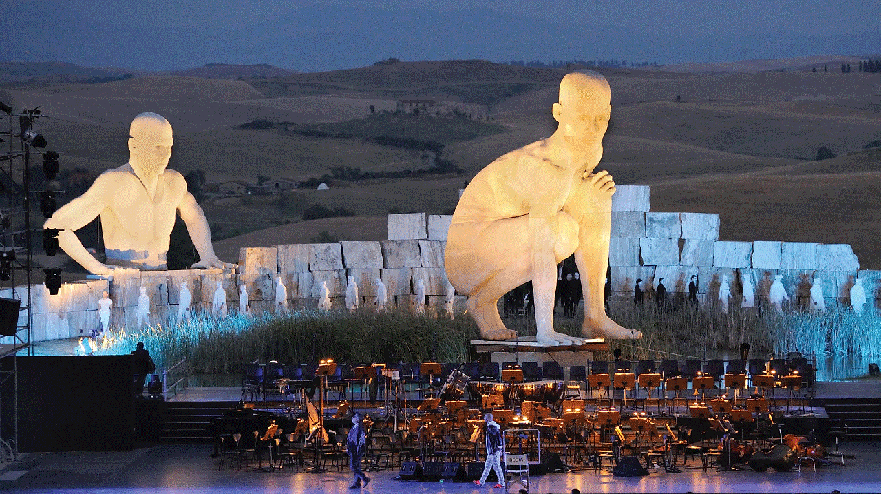 Two monumental white figures above the stone stage at Teatro del Silenzio at dusk, with the orchestra's chairs set out below