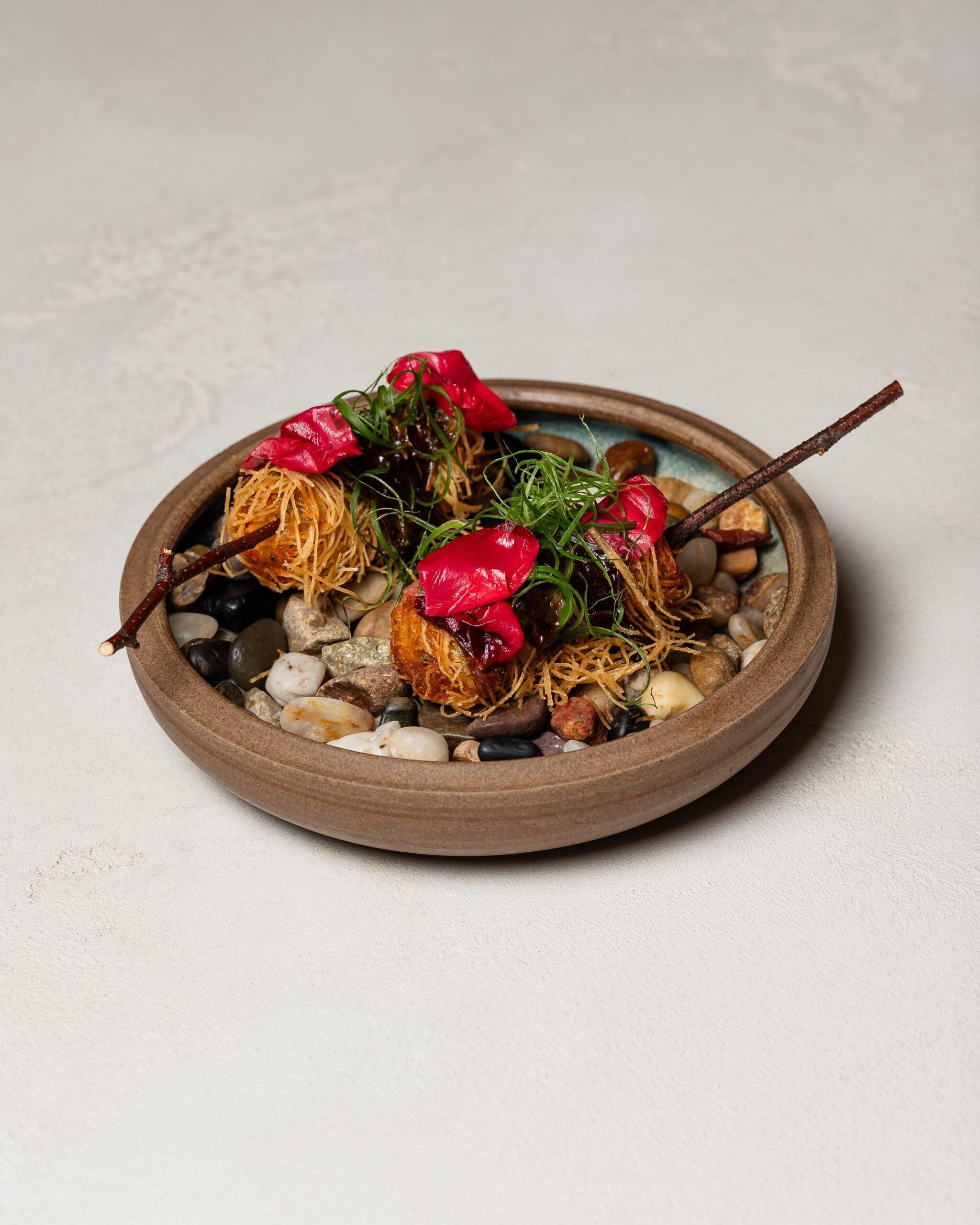 A HIDE course in a stoneware bowl over smooth pebbles, topped with crisp pastry, herbs and red petals