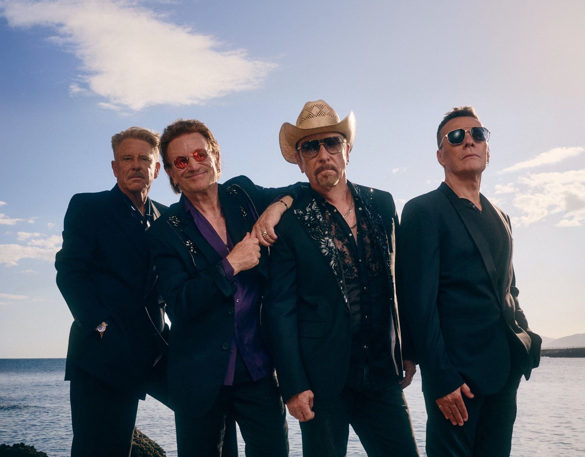 U2, the four band members in dark suits, standing by the sea under a blue sky