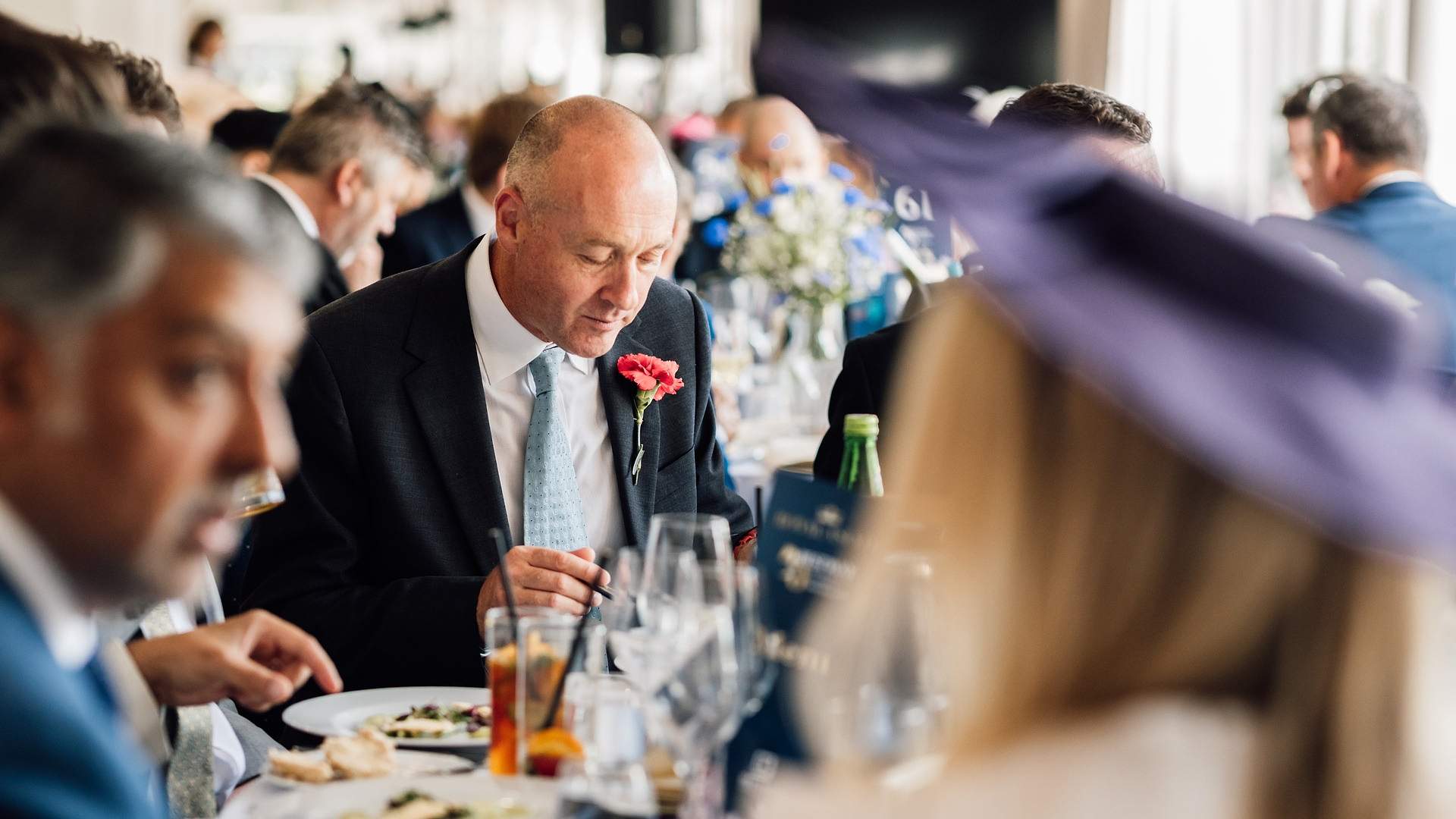 A guest in navy morning dress with a carnation buttonhole dining indoors at Royal Ascot