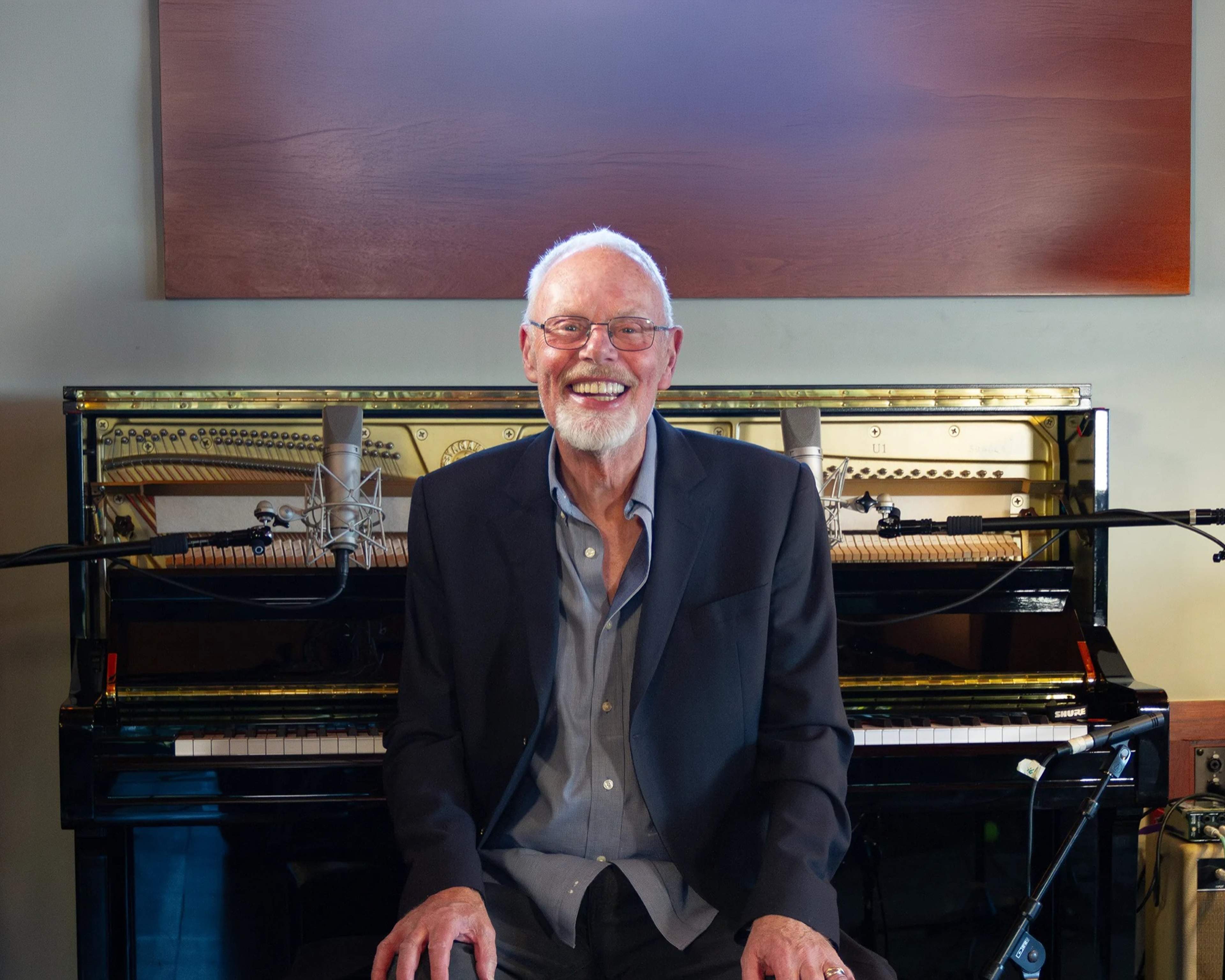 Bob Harris smiling at an upright piano in a recording studio, with microphones set up around it