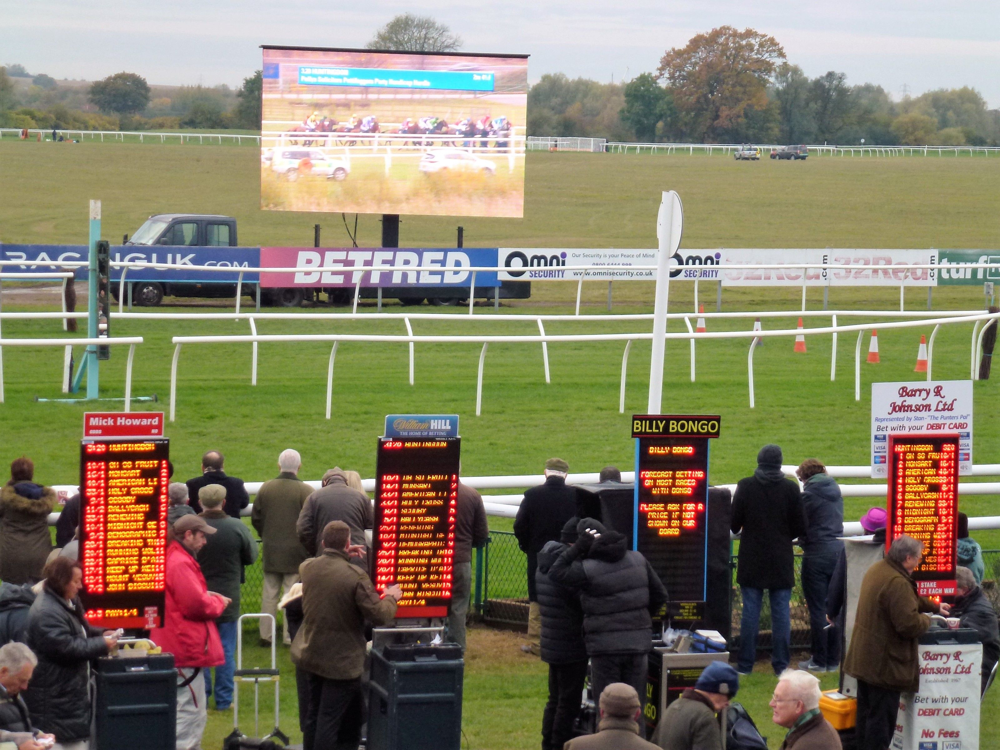 Four on-course bookmakers at their pitches with electronic odds boards, and racegoers standing in front of them