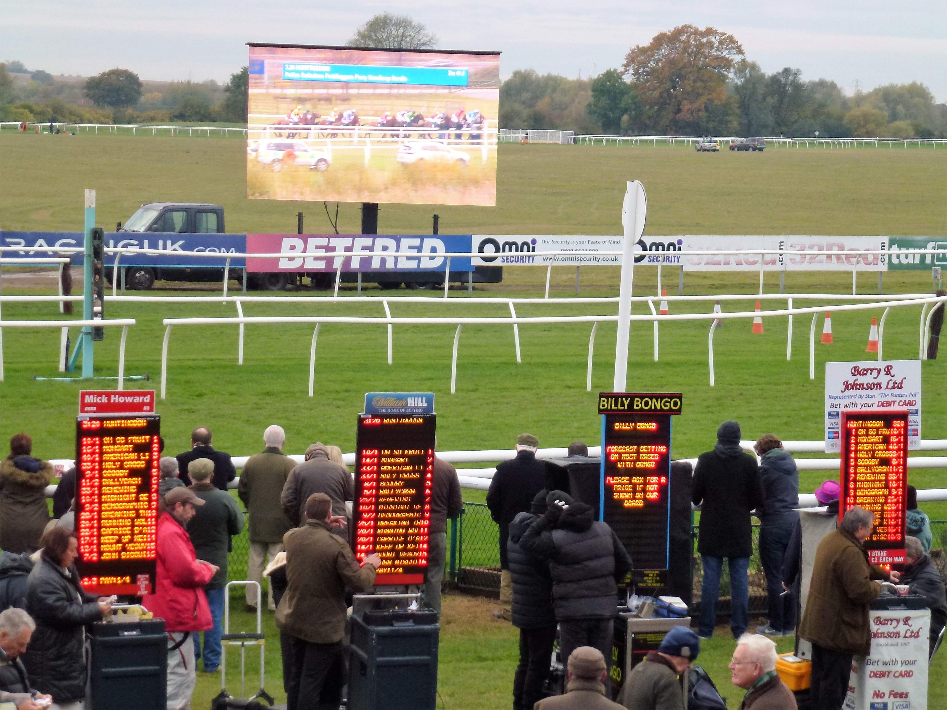 Four on-course bookmakers at their pitches with electronic odds boards, and racegoers standing in front of them