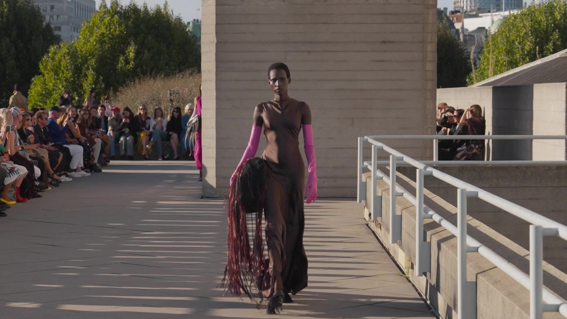 A model in a brown gown and pink gloves walks the outdoor runway in afternoon sun at Roksanda's September 2026 show