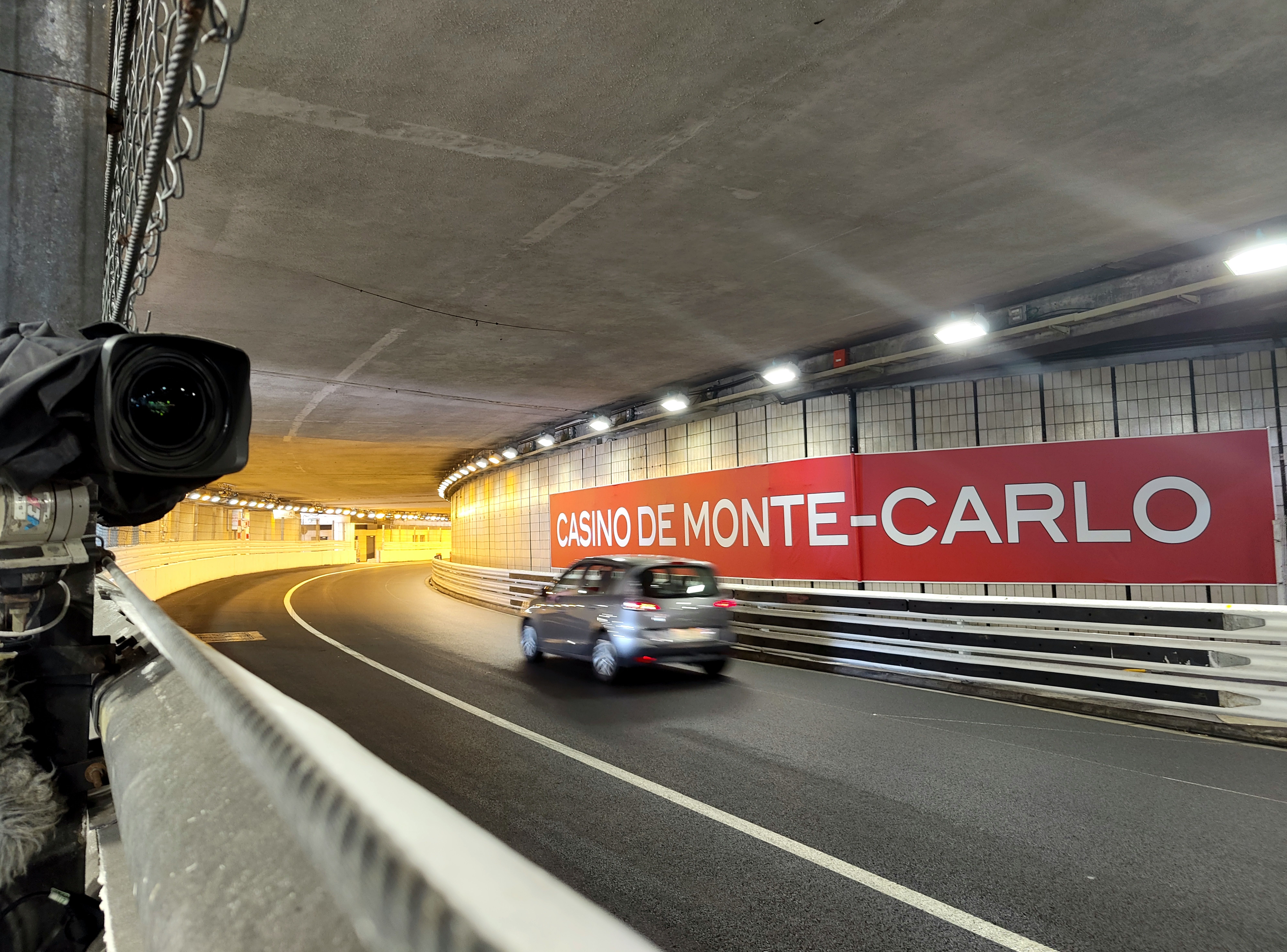 A car driving through the Monaco tunnel, with race barriers and a trackside camera along the inside wall
