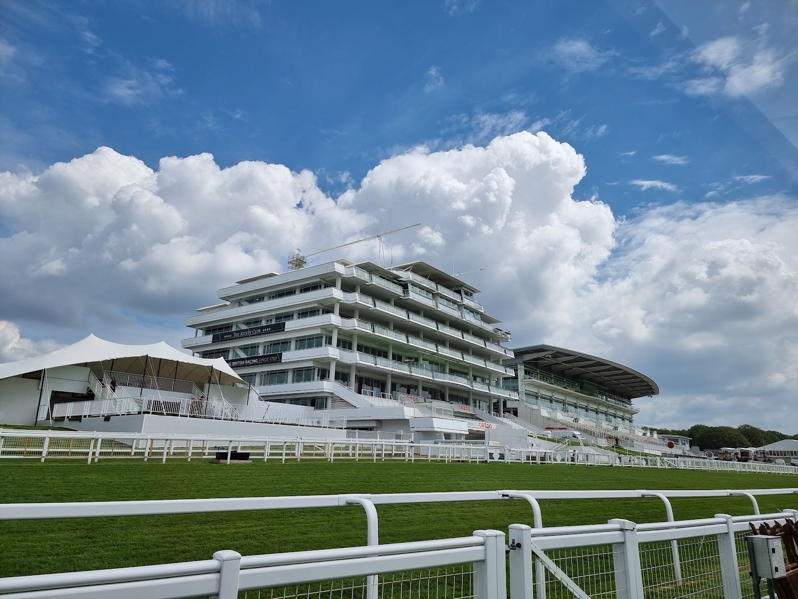 The Queen Elizabeth II Stand at Epsom Downs, a white tiered grandstand behind the running rail, with a marquee to its left and the Grandstand to its right under a blue sky