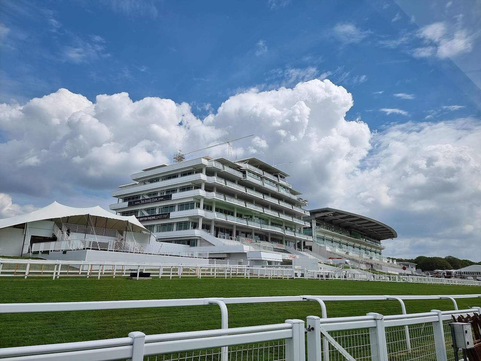 The Queen Elizabeth II Stand at Epsom Downs, a white tiered grandstand behind the running rail, with a marquee to its left and the Grandstand to its right under a blue sky