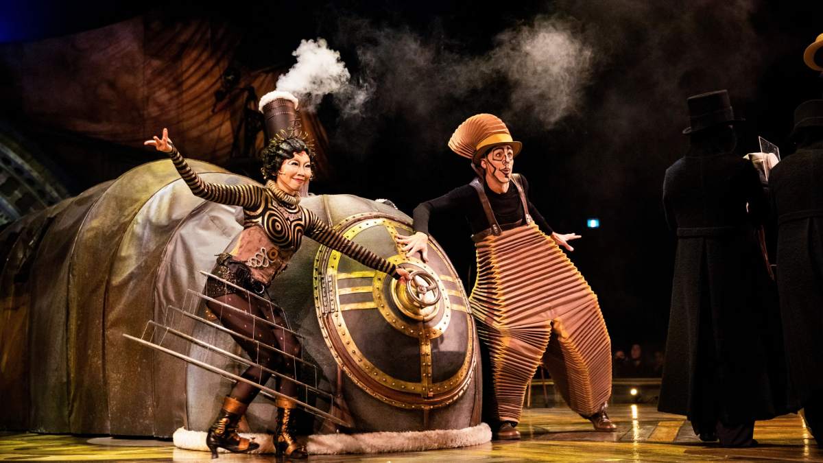 Two KURIOS characters beside a steam-powered machine on stage