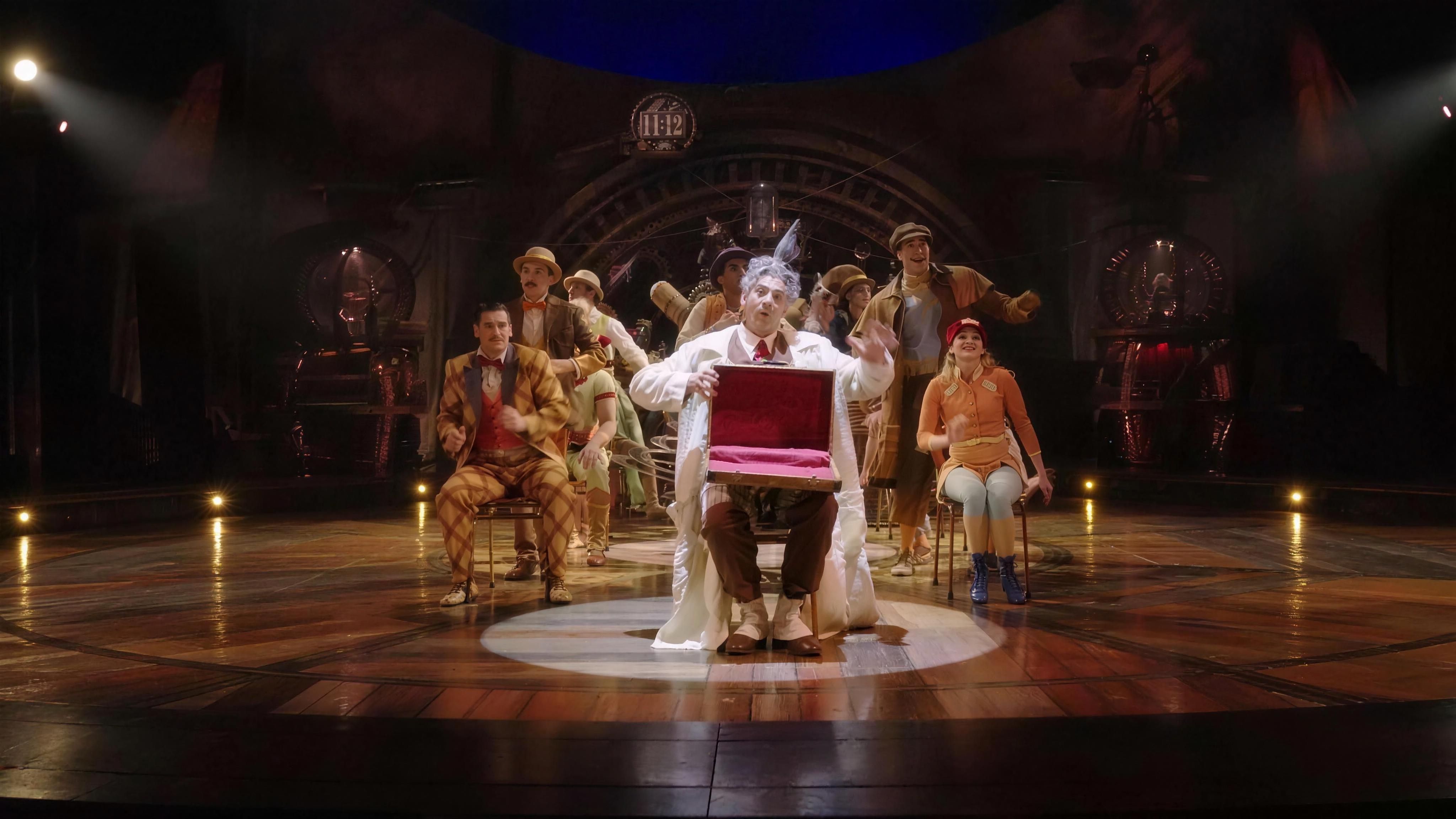 The Seeker holds up a red case at the centre of the KURIOS stage, the cast gathered behind him