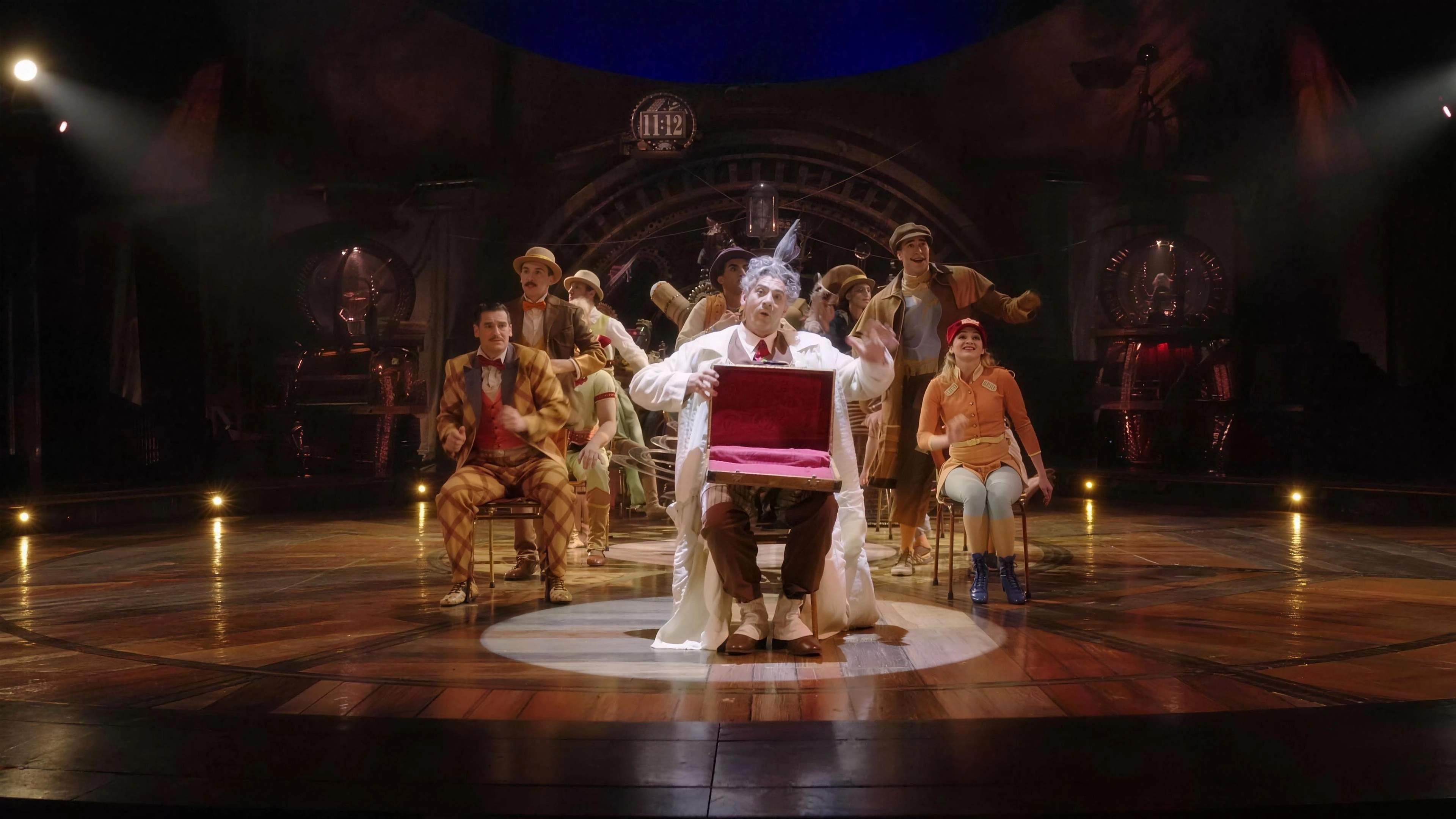 The Seeker holds up a red case at the centre of the KURIOS stage, the cast gathered behind him