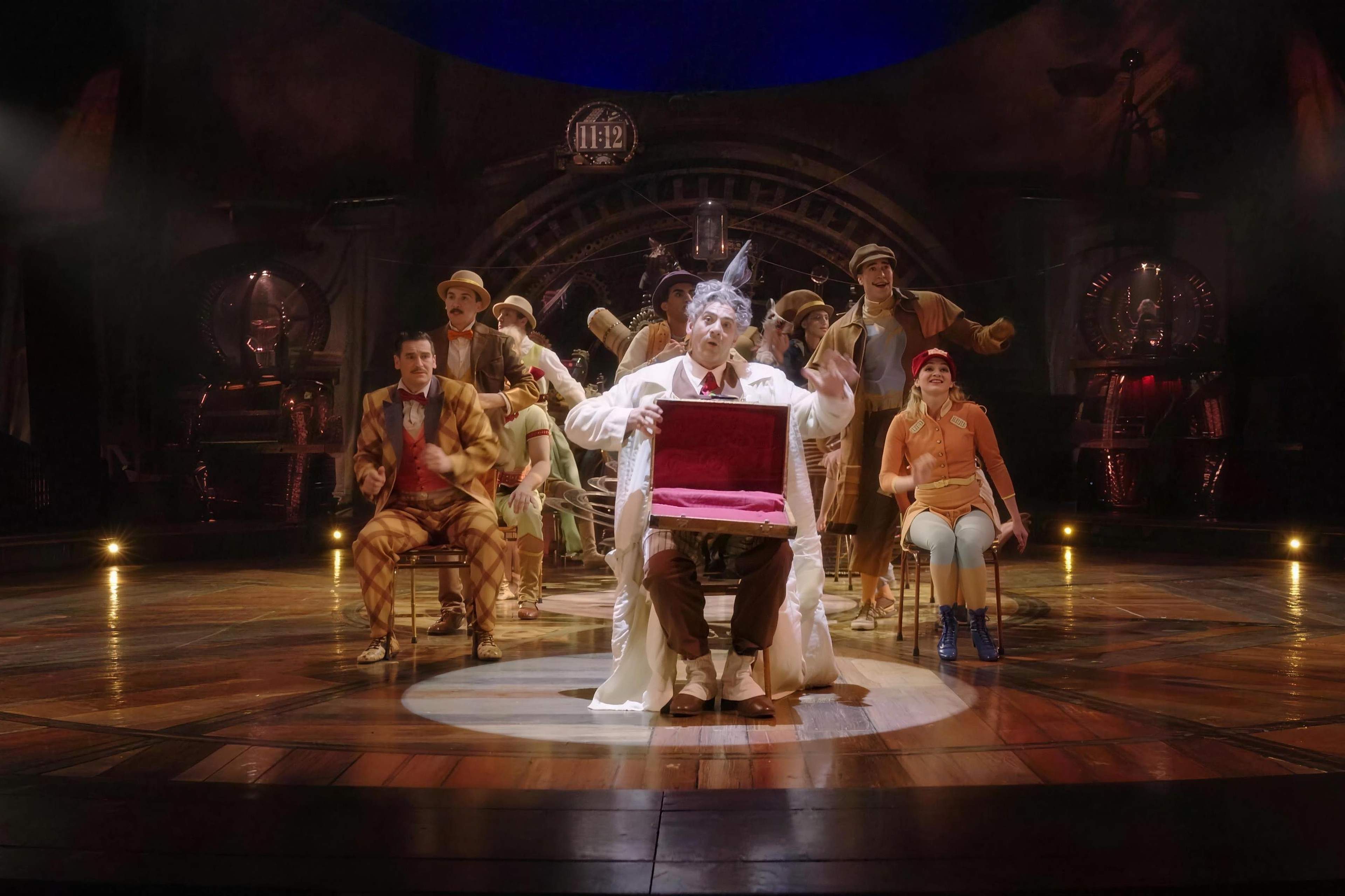 The Seeker holds up a red case at the centre of the KURIOS stage, the cast gathered behind him