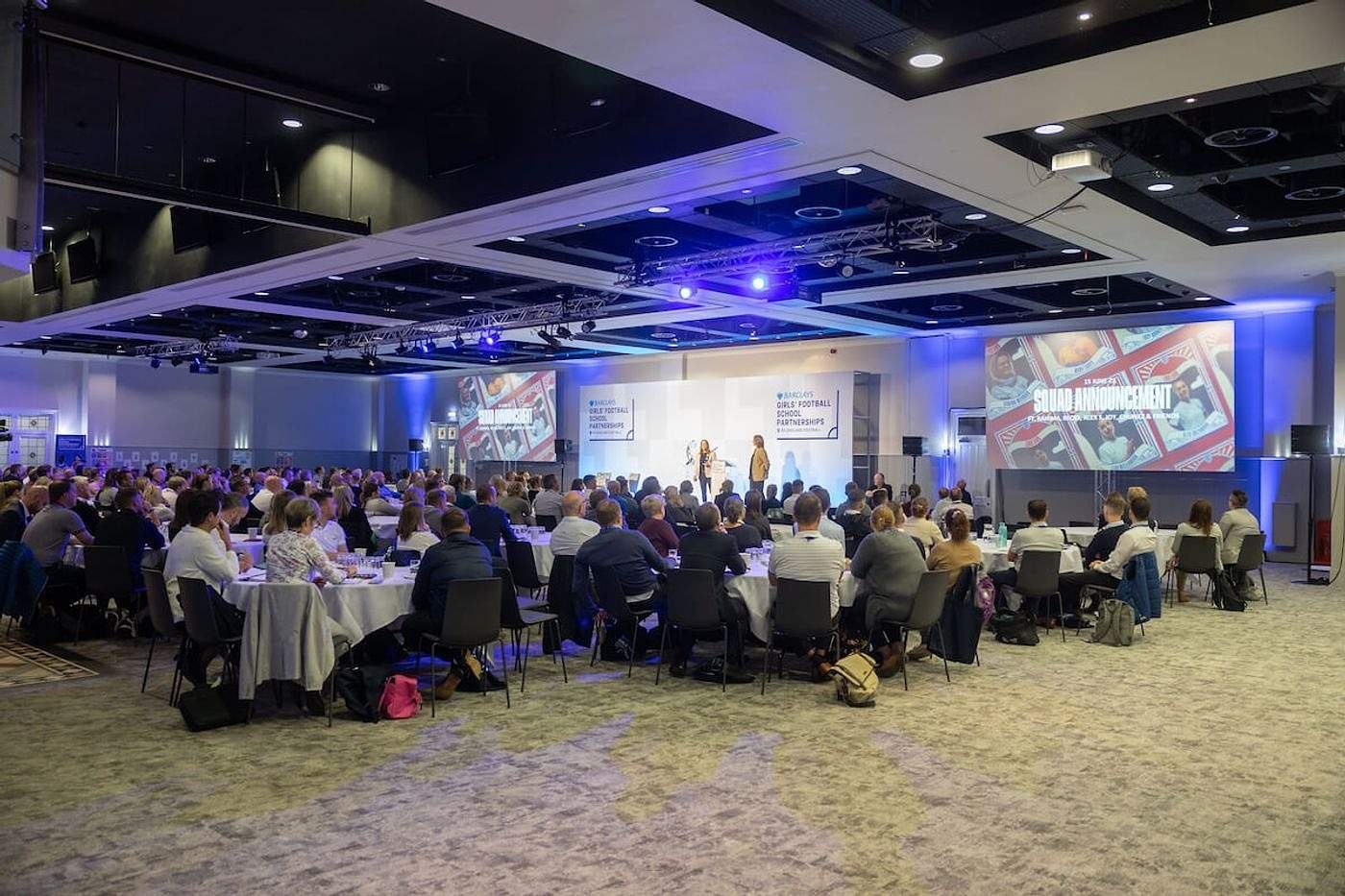 Guests at round tables facing a stage with screens under blue lighting in the Lower Grounds suite at Villa Park