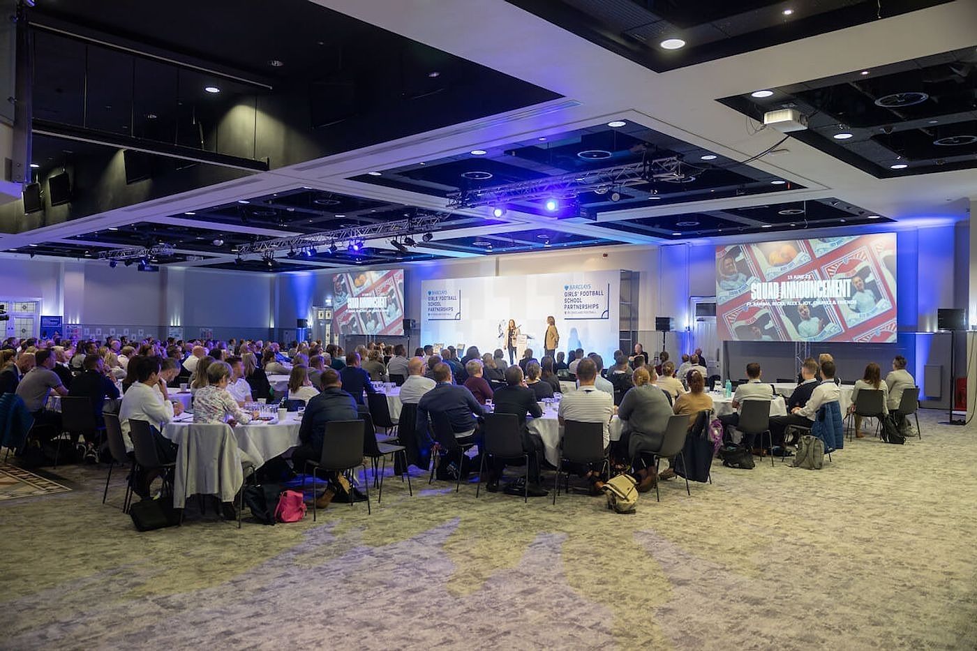 Guests at round tables facing a stage with screens under blue lighting in the Lower Grounds suite at Villa Park