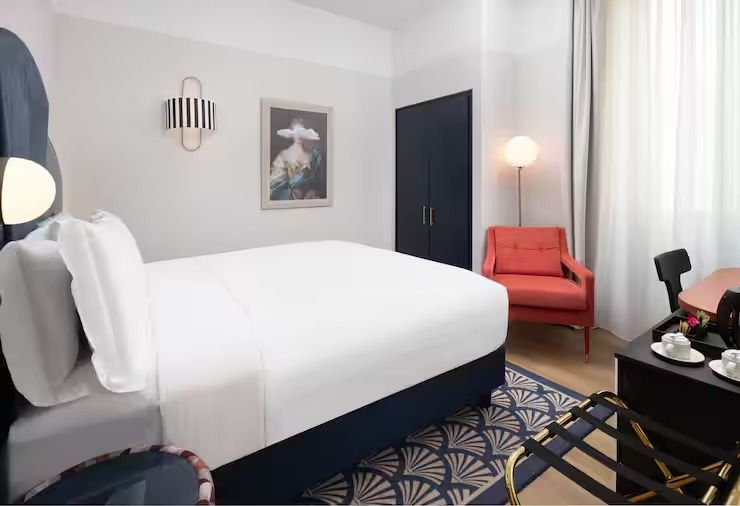 Bed dressed in white against a navy base, below a framed portrait and a striped wall light, with a coral armchair and a fan-pattern rug